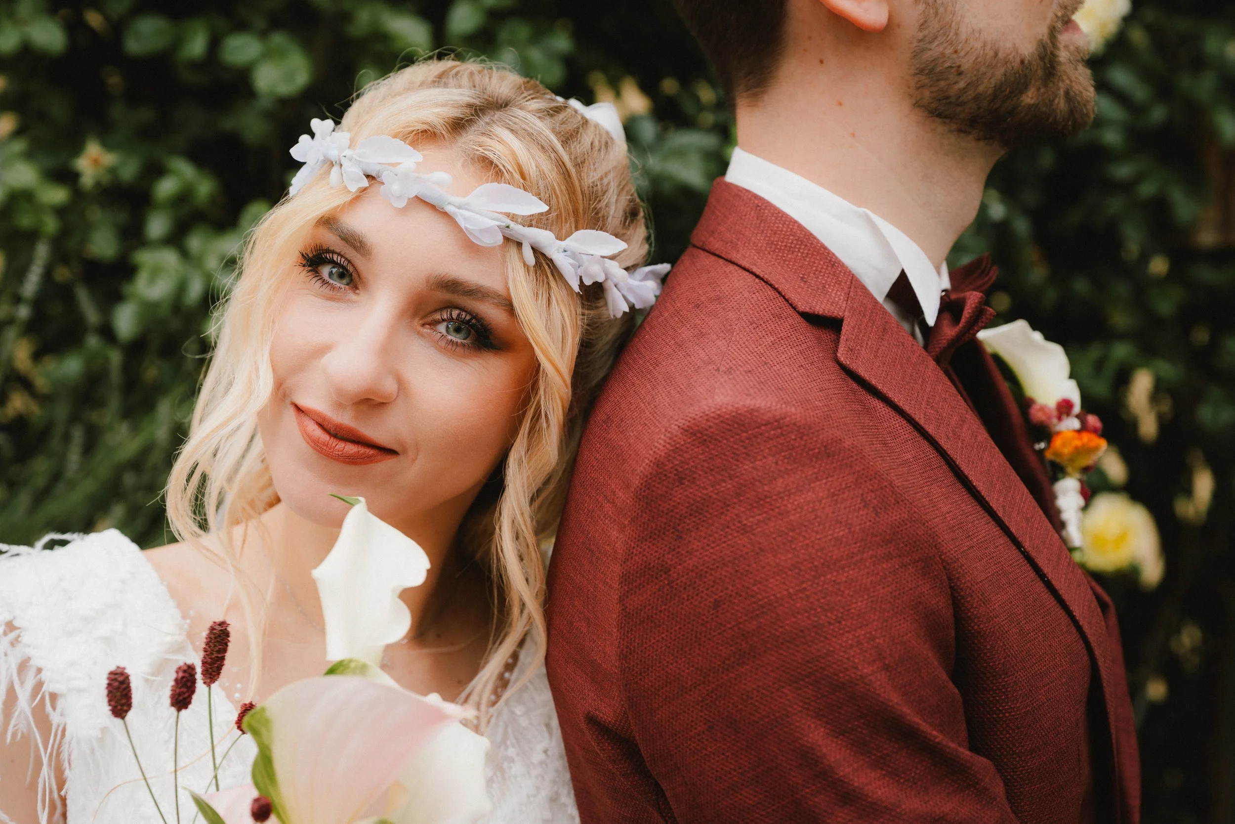 Ein Brautpaar steht im Grünen, die Braut lehnt ihren Kopf an die Schulter des Bräutigams, trägt einen weißen Brautkleid mit Spitze und eine weiße Blumenkrone, während der Bräutigam einen roten Anzug mit weißen Hemd und Fliege trägt.