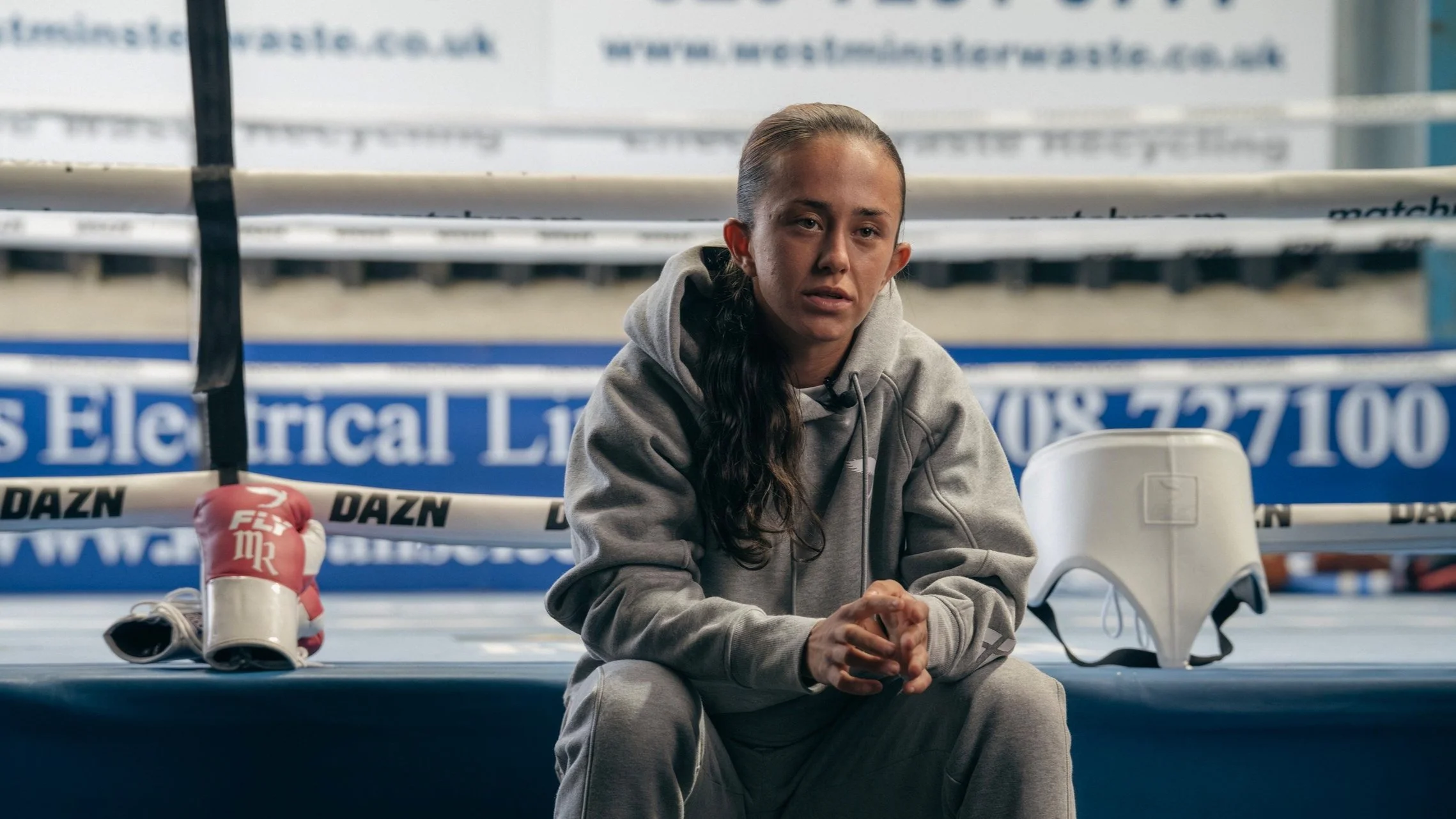 A young woman sitting in a boxing gym, wearing a grey hoodie and sweatpants, with boxing gloves and a face shield on the floor nearby. She appears tired or stressed, with a boxing ring in the background.
