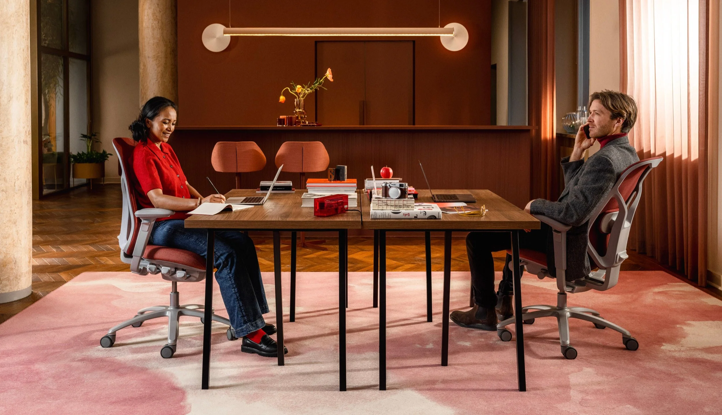 A woman and a man face each other sitting in Mero Meesh chairs by designers at Fjaryl and Flokk. In a room with red and pink tints
