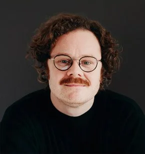 Person med brunt, krøllet hår, briller og skæg, iført sort T-shirt, mod en mørk baggrund.