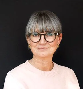 Et portrætfoto af en ældre kvinde med gråt hår, briller og hvid sweater mod en sort baggrund.