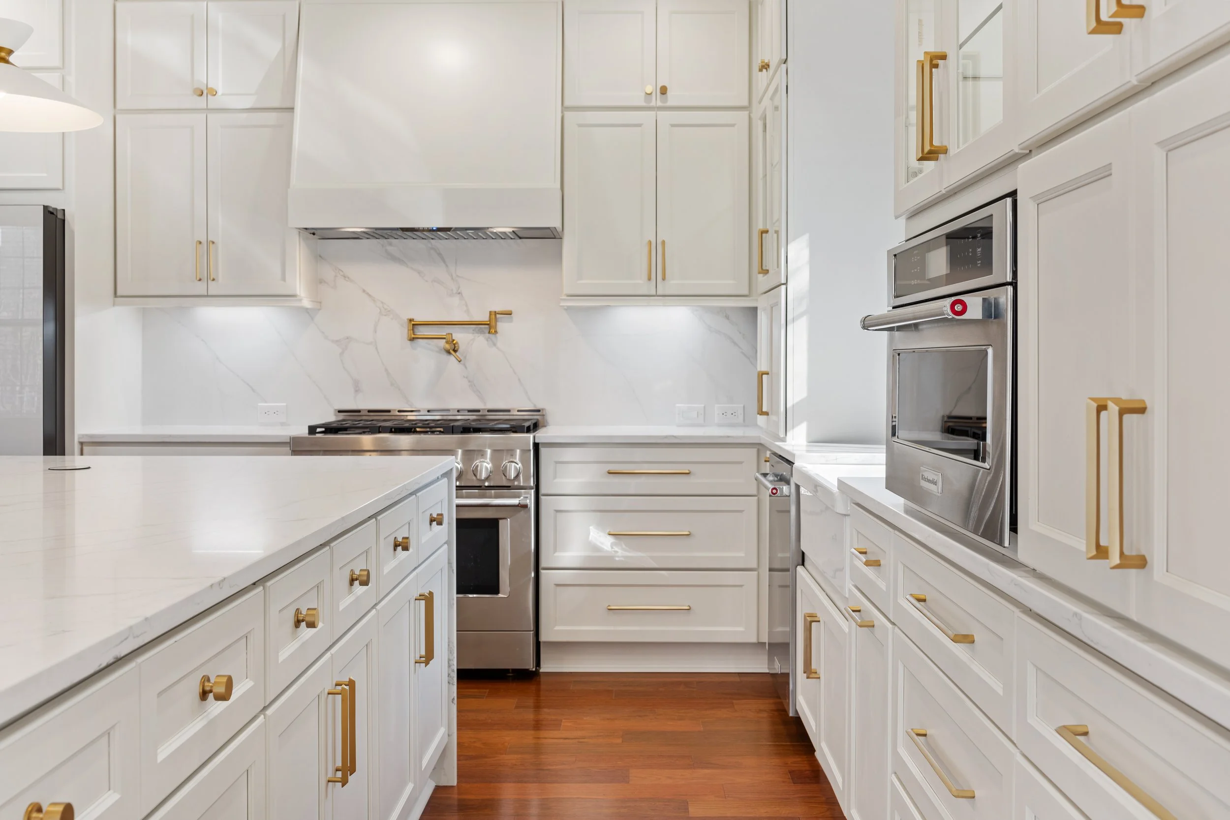 Modern white kitchen with gold hardware, stainless steel appliances, and a marble backsplash. Features kitchen cabinets, oven, and a central island with a smooth white countertop.