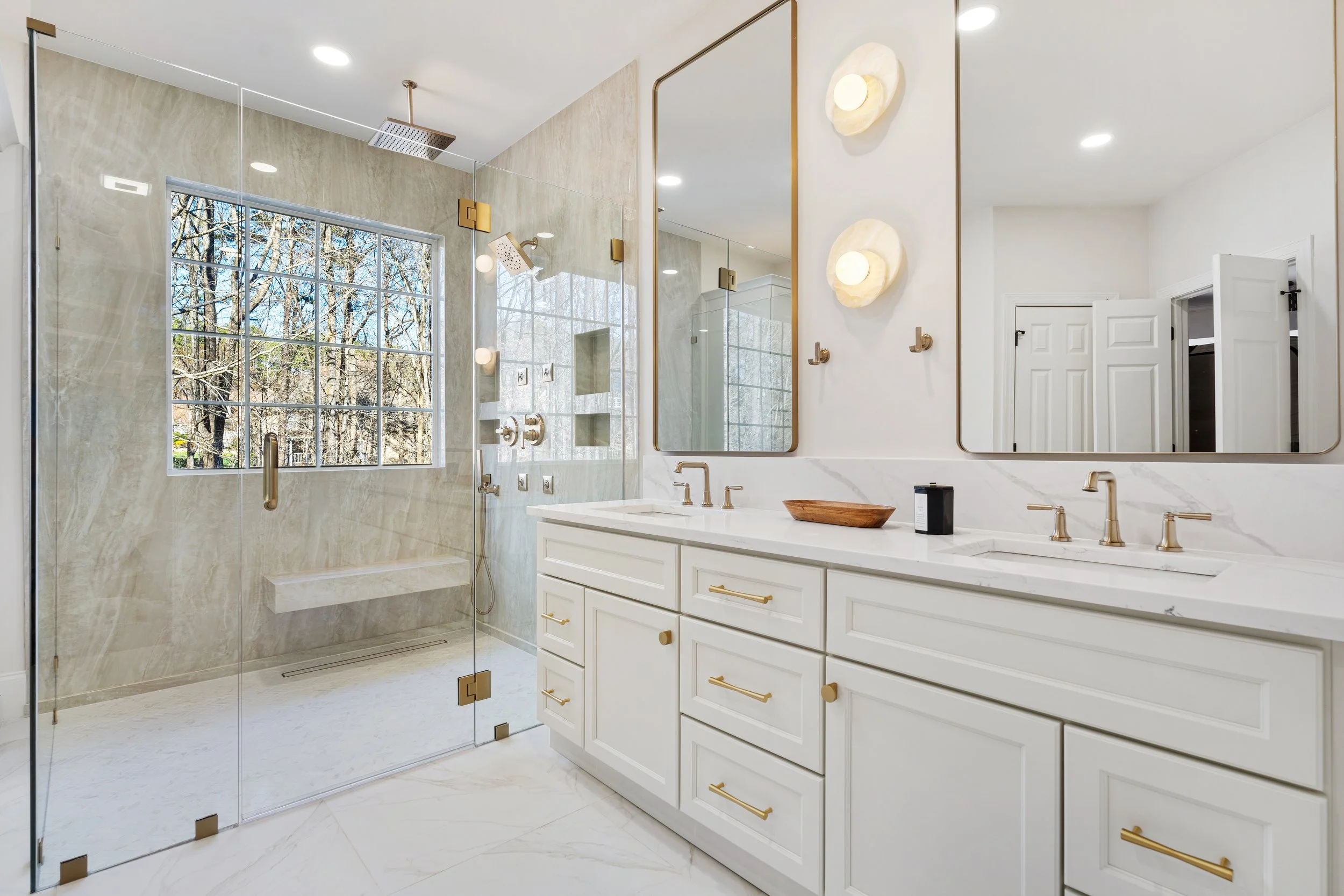 Modern bathroom with a large glass shower enclosure, white double vanity, and gold hardware, including two mirrors, wall-mounted lights, and a window overlooking trees outside.