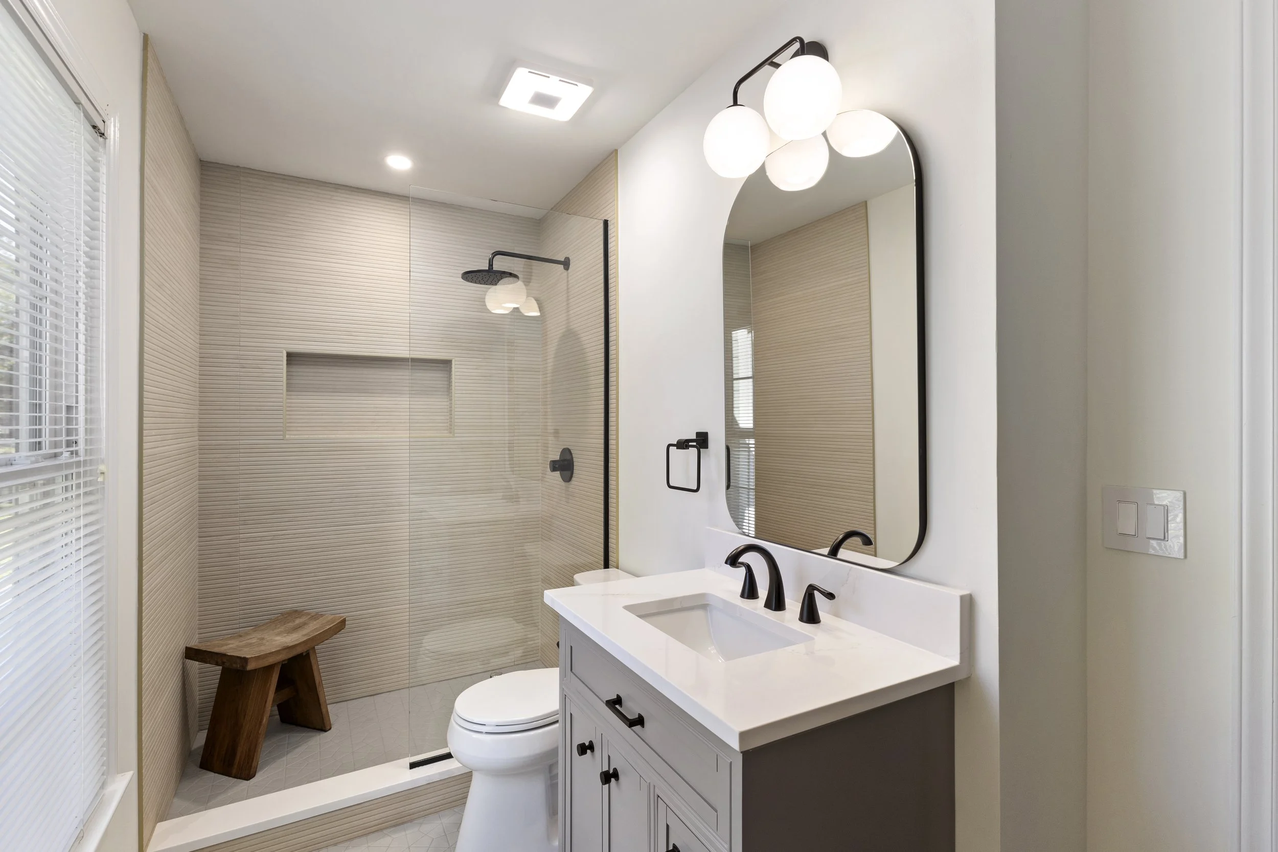 Modern bathroom with glass-enclosed shower, wooden bench, white vanity with black fixtures, large mirror, and a window with blinds.