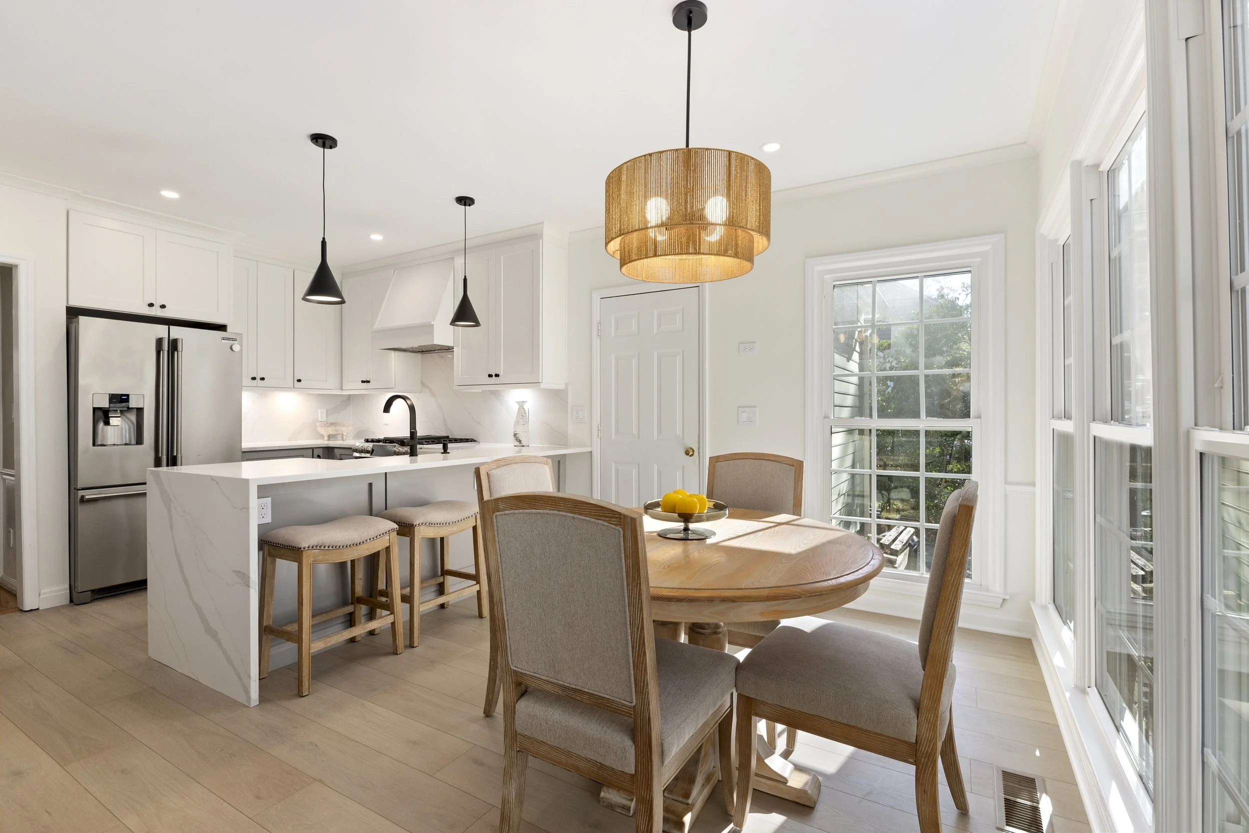 Bright kitchen and dining area with white cabinets, stainless steel refrigerator, island with seating, wooden dining table with four chairs, large windows, and a hanging light fixture.
