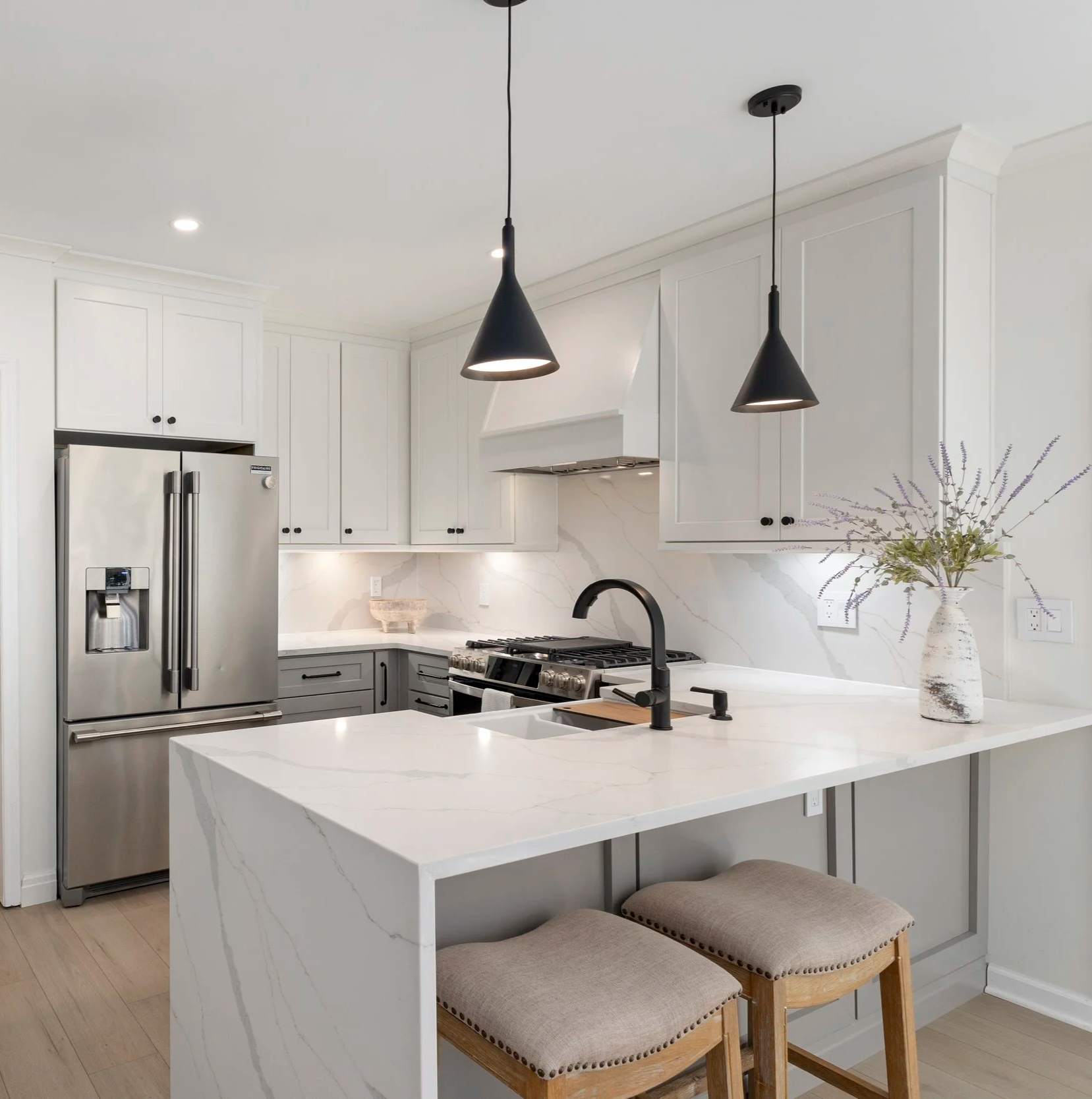 Modern white kitchen with black fixtures, stainless steel refrigerator, and a marble island with two beige stools and a vase with flowers.