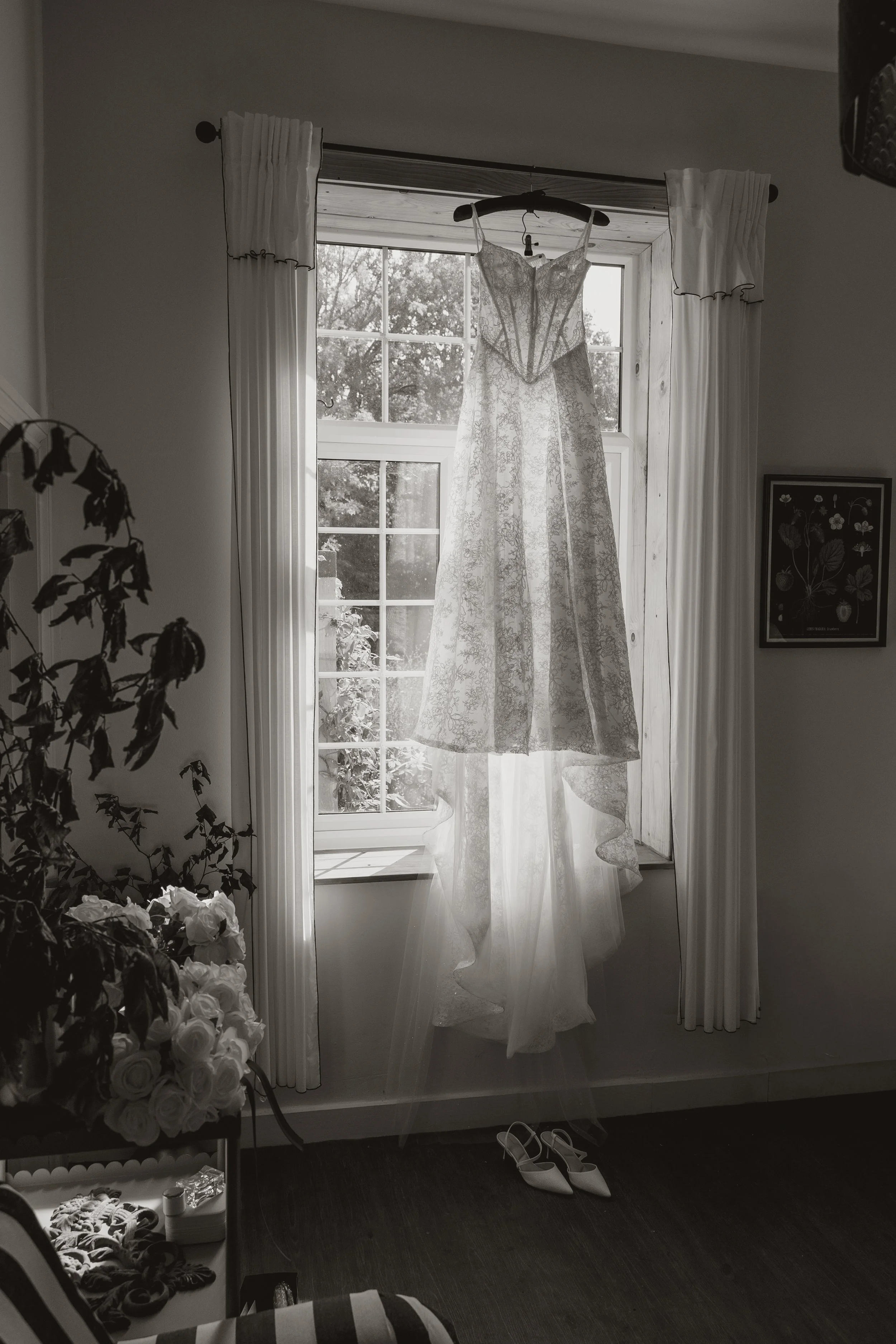 A wedding dress hanging on a hanger in front of a window with white curtains, surrounded by flowers and shoes on the floor.