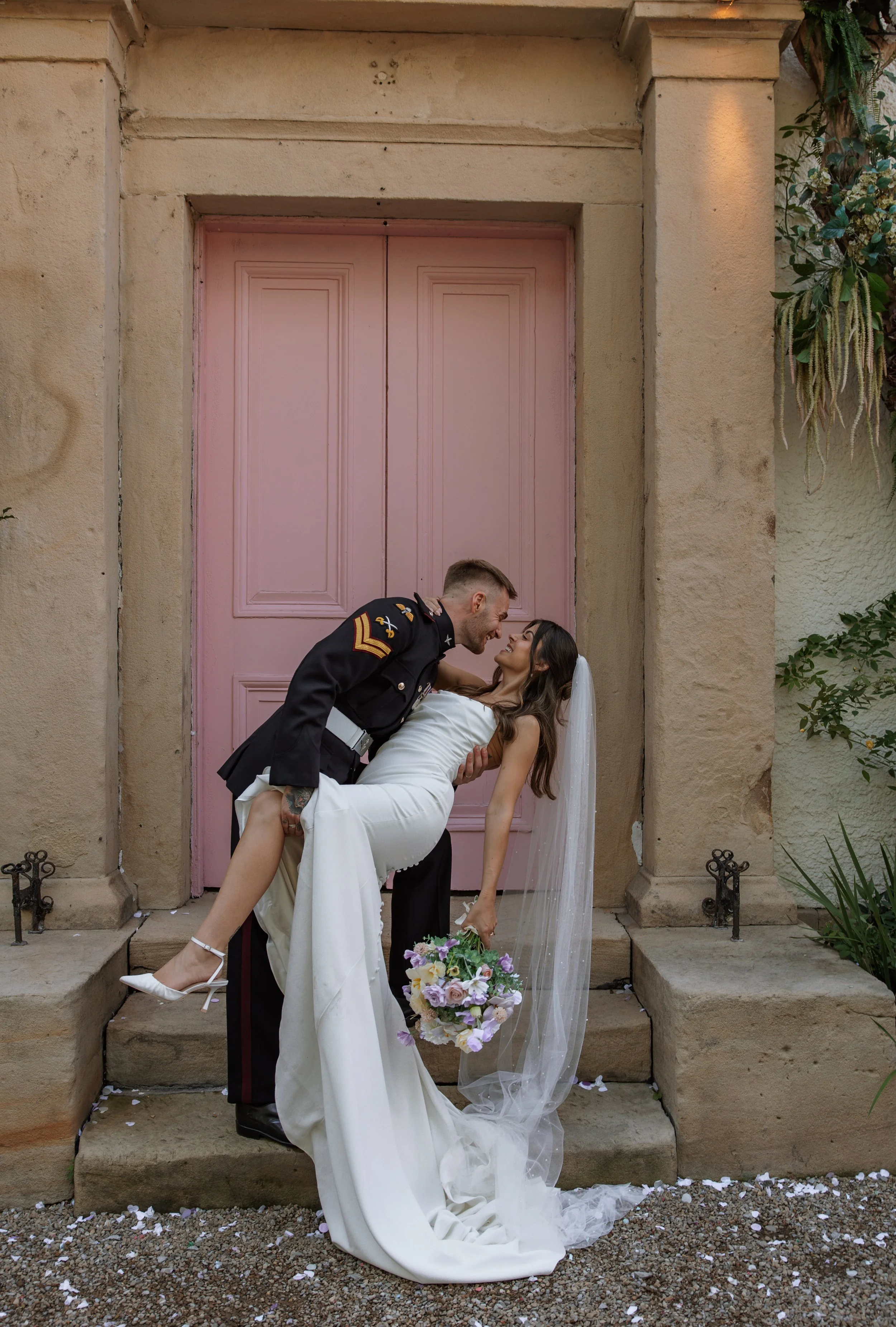 A bride in a white wedding gown and veil being held by a groom in a military uniform, standing on steps in front of a pink door, with scattered flower petals on the ground.