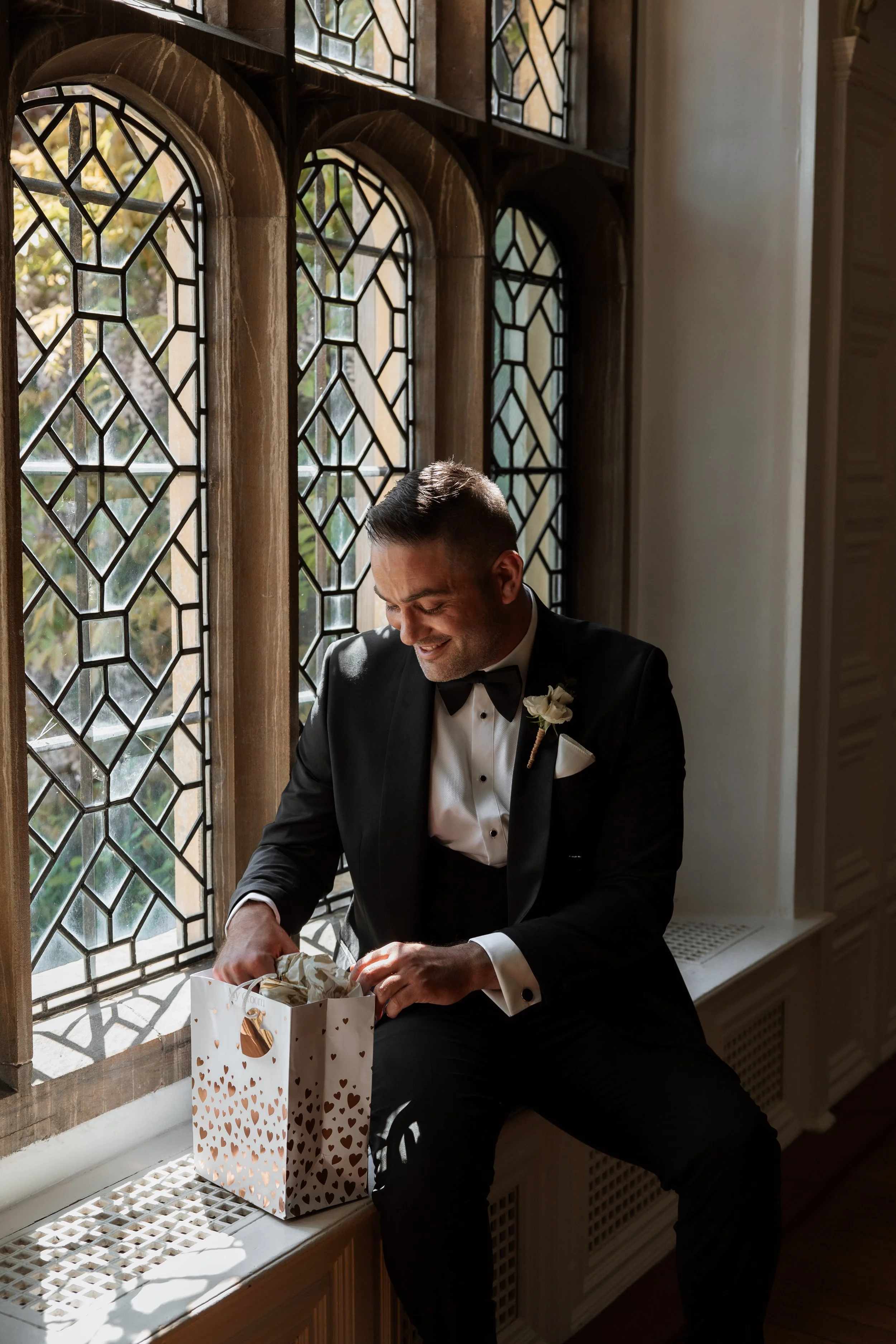 A man in a tuxedo opening a gift bag near a stained glass window.
