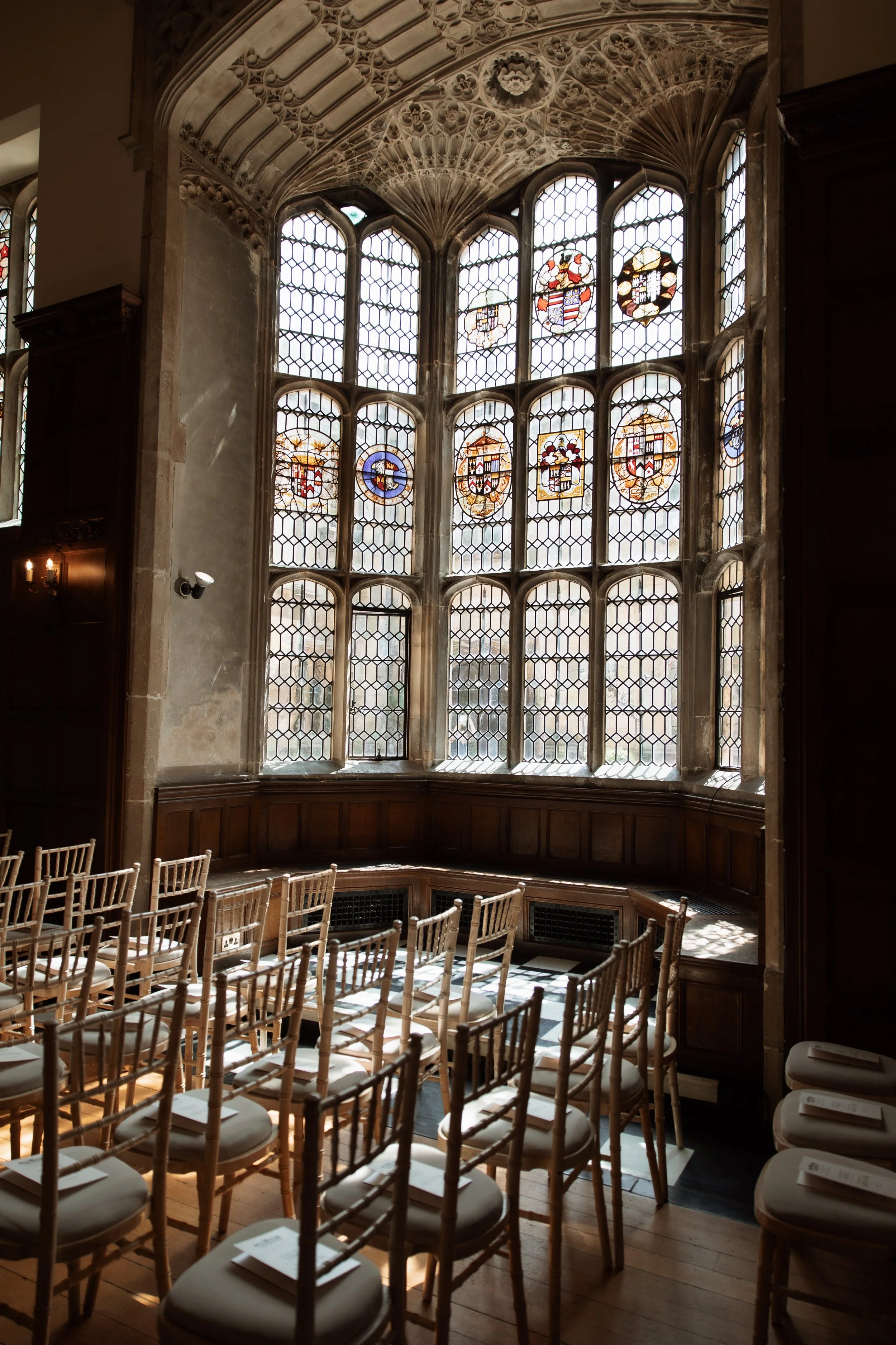 A room with a large, ornate stained glass window featuring coats of arms and heraldic symbols, surrounded by detailed stonework. Wooden chairs are arranged in rows facing towards the window, with some papers on the seats.