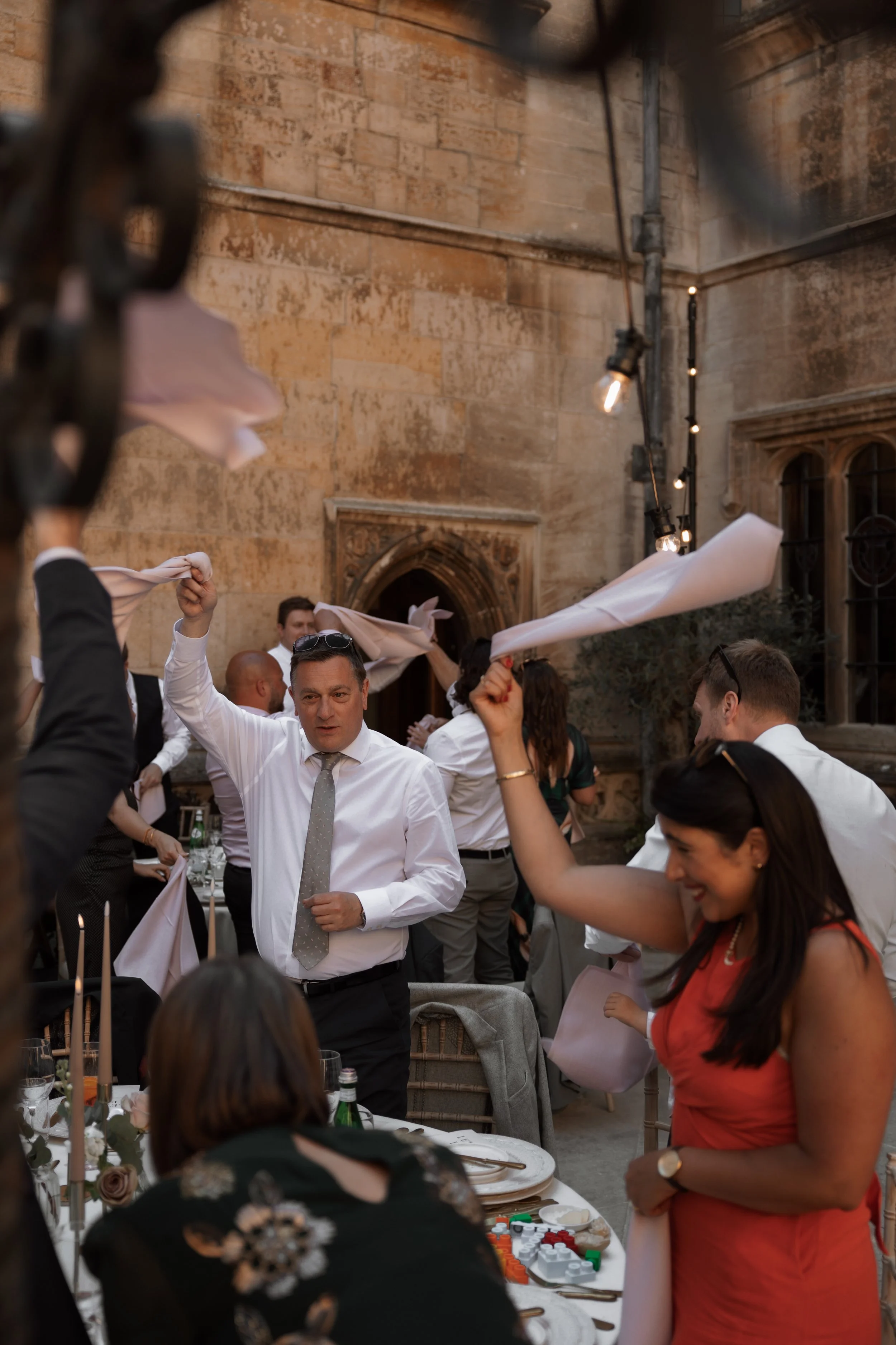 People celebrating at an outdoor event with white napkins in the air, seated and standing around a table, in a historic stone courtyard with warm lighting.