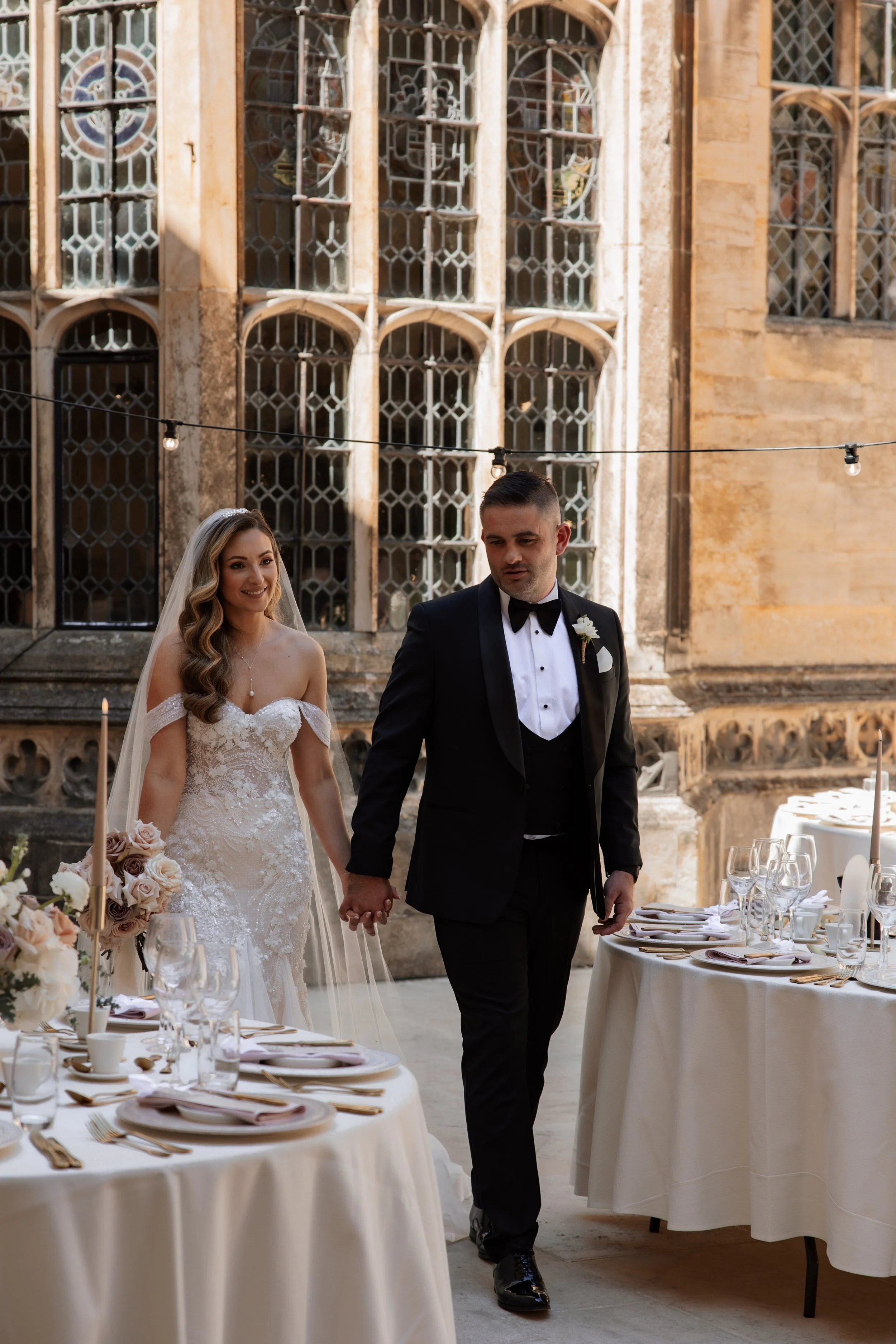 A bride and groom walking hand in hand at their wedding reception outdoors, with decorated tables and the historic chapel windows in the background.
