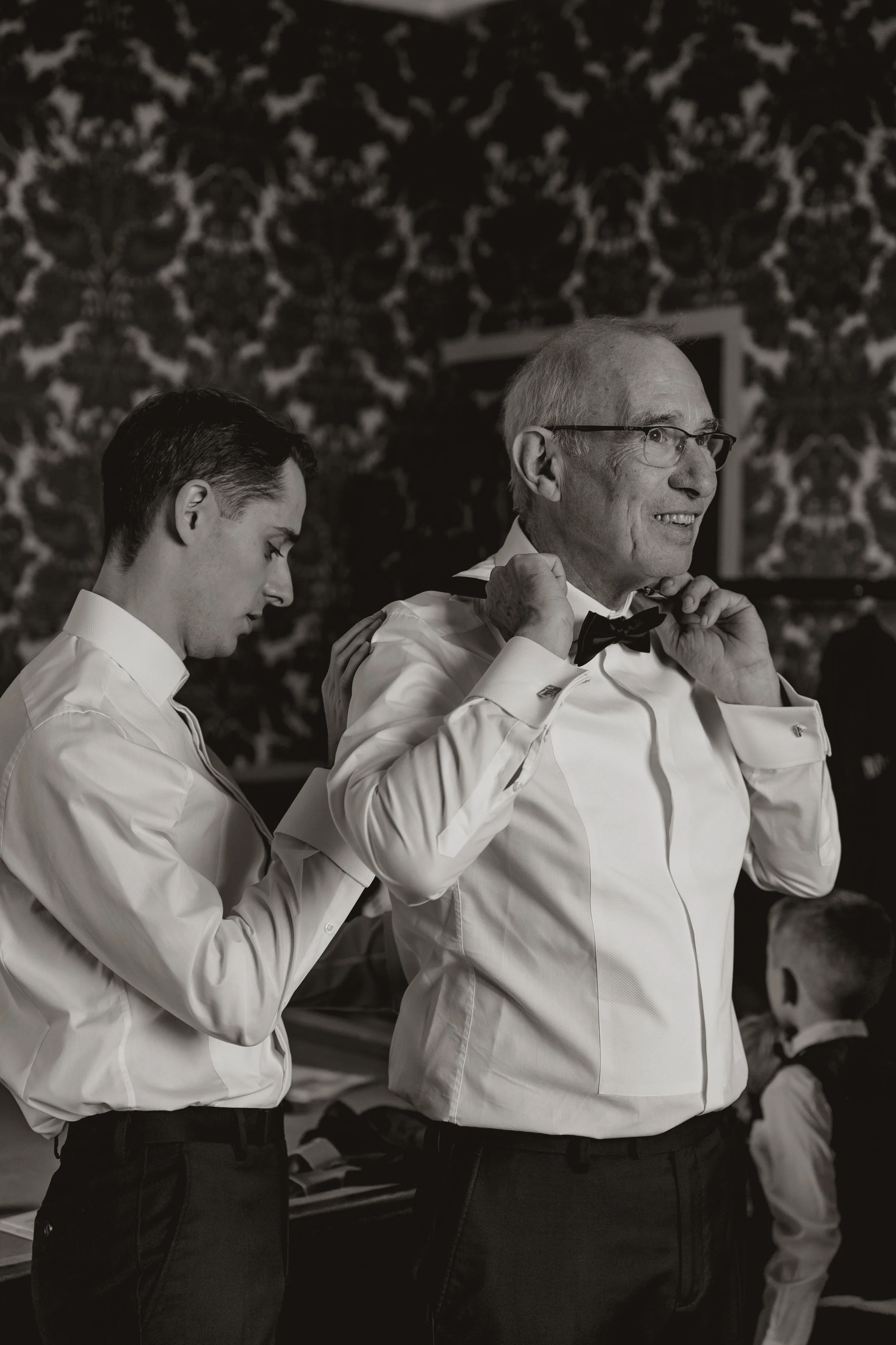An elderly man with glasses helping a young man adjust a bow tie in a well-decorated room.