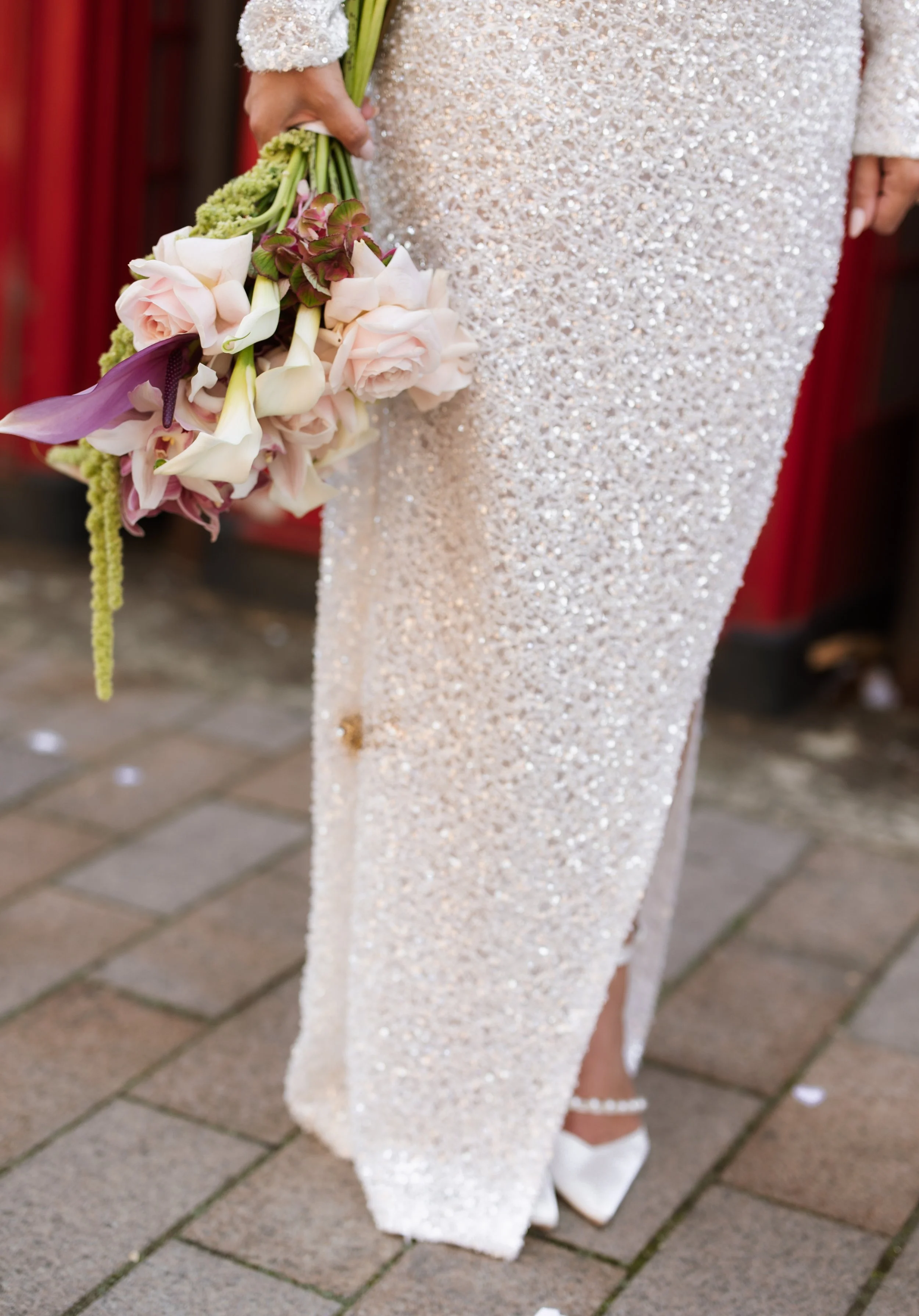 Close-up of a bride holding a bouquet of light pink roses, white calla lilies, and greenery, wearing a sequined white wedding dress and white high heels, standing on a brick pathway outdoors.