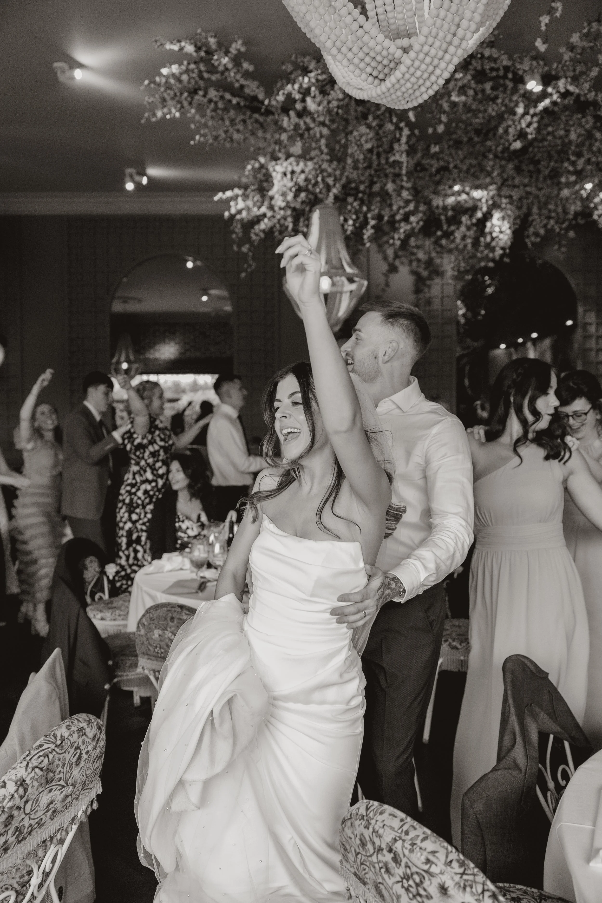 Black and white photo of a joyful wedding reception with people dancing, including a woman in a wedding dress and a man in a white shirt dancing closely, with a decorated ceiling and hanging floral arrangements.