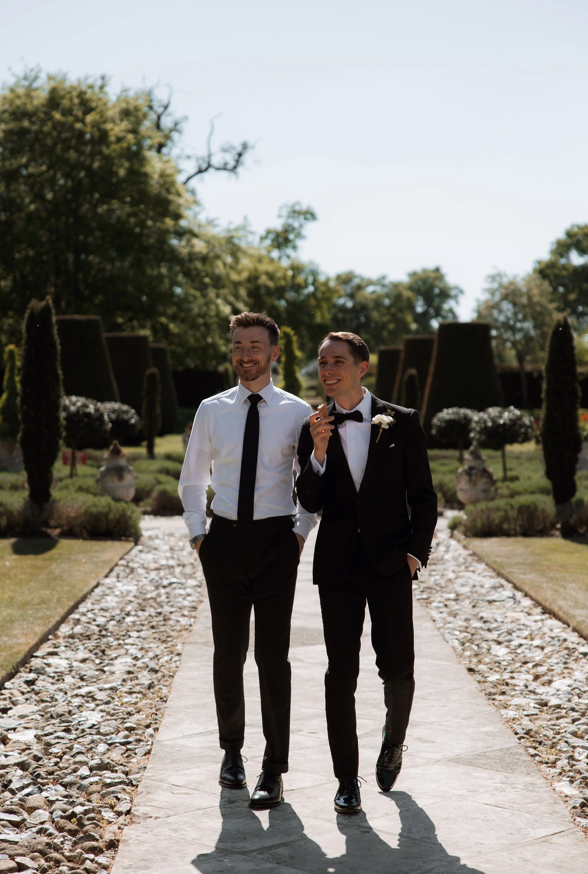 Two men in formal attire walking outdoors, one in a tuxedo and the other in a white shirt and black tie, smiling and enjoying a sunny day in a landscaped garden.