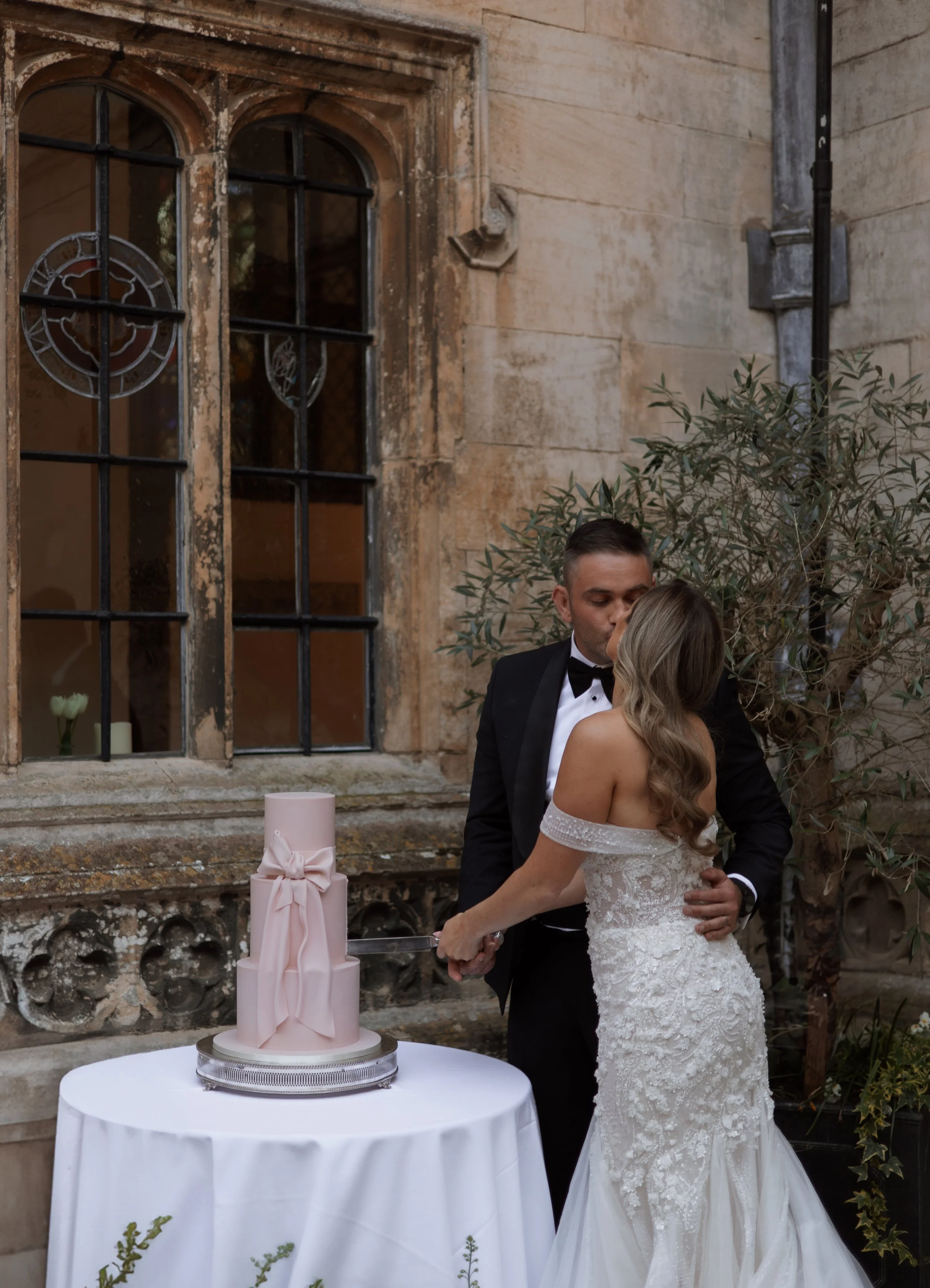 A bride and groom cut a pink wedding cake together at their wedding reception outdoors, with a stone building and an olive tree in the background.