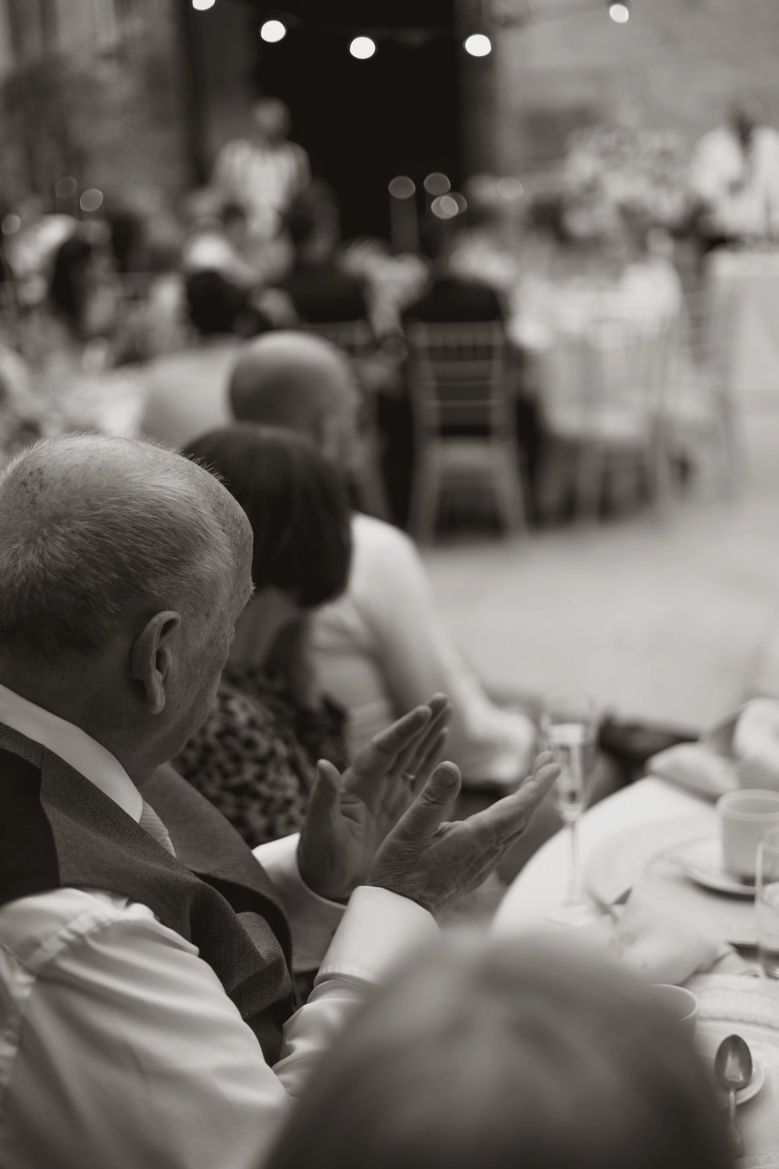 A black-and-white photo of an elderly man with short white hair clapping his hands at a formal gathering, with other seated guests and a speaker or presenter in the background.