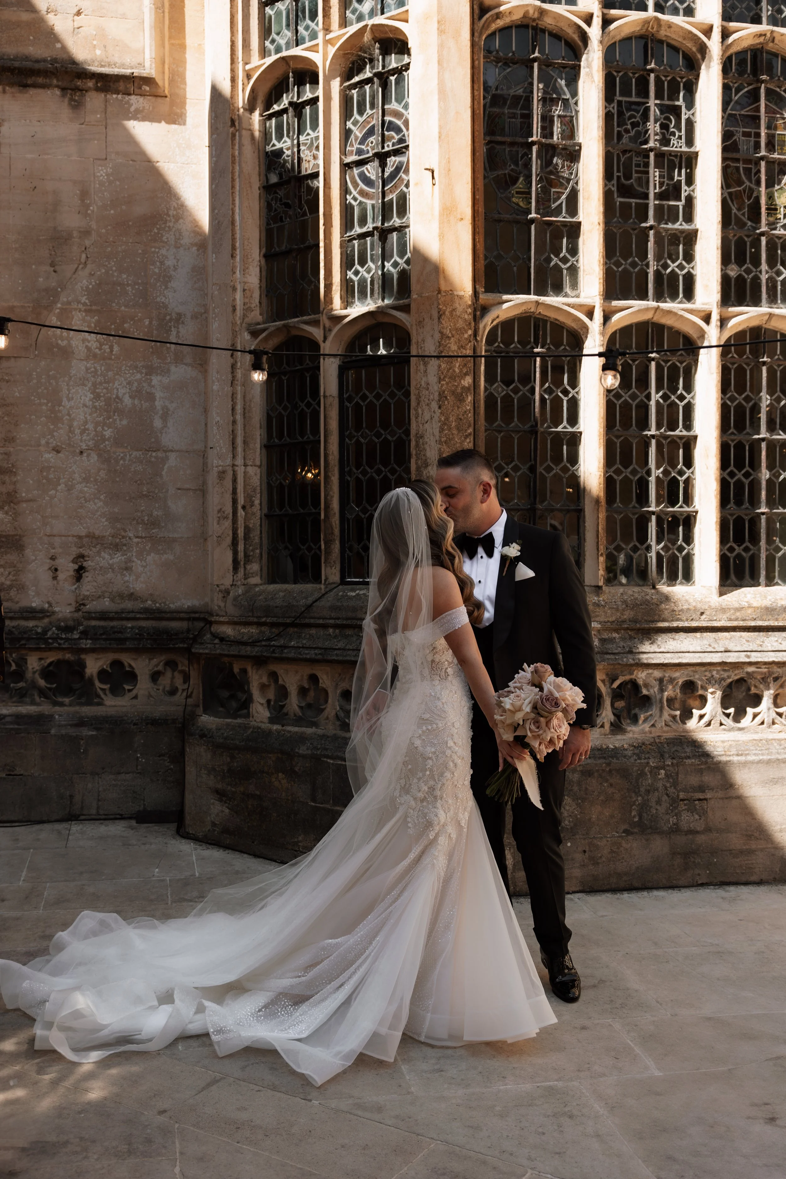 A bride and groom share a kiss outdoors in front of a historic stone building with stained glass windows. The bride wears a white wedding gown with a long train and veil, holding a bouquet of roses. The groom is dressed in a black tuxedo with a bow t