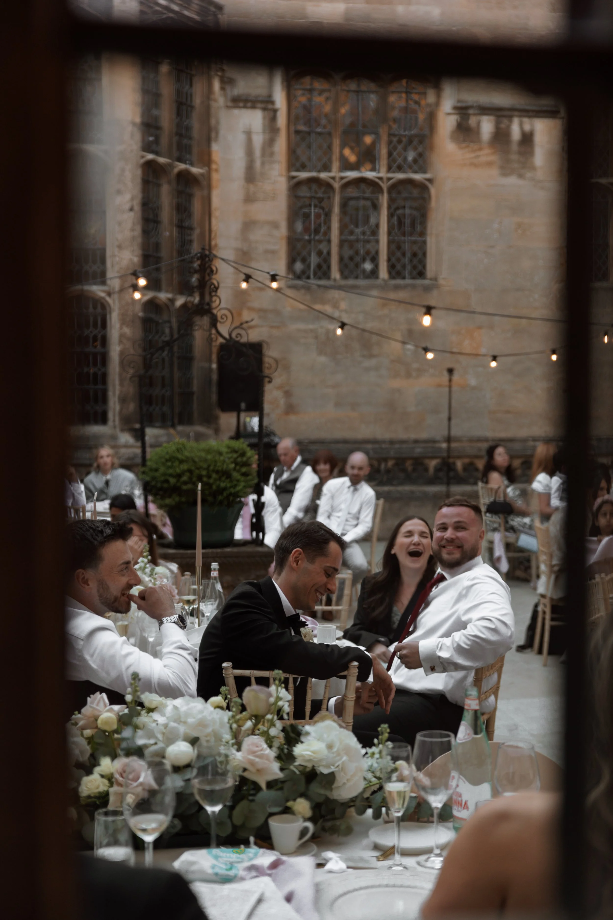 People laughing and enjoying a celebration at a wedding reception in an outdoor courtyard with hanging string lights and large historical windows in the background.