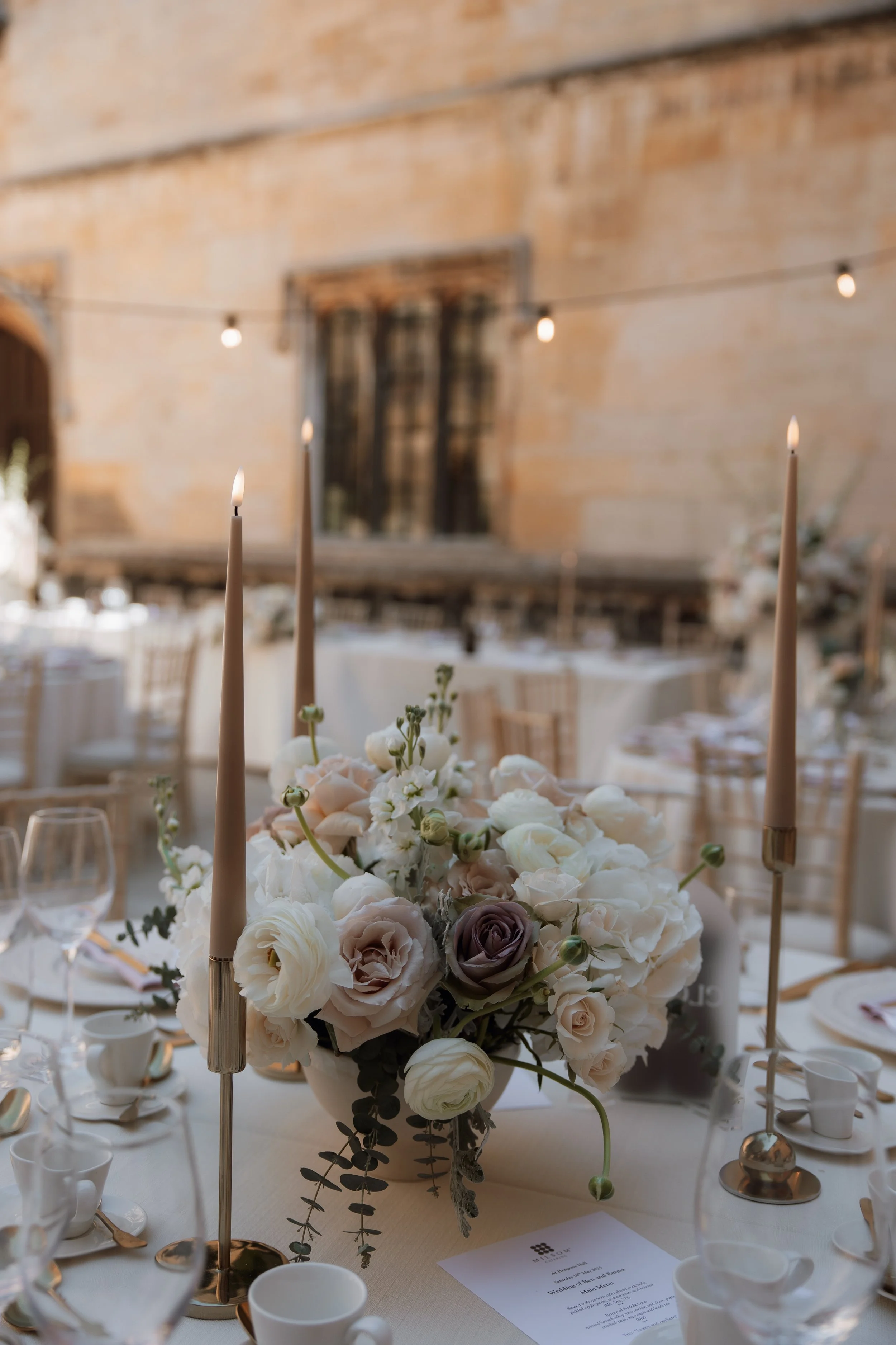 Elegant table centerpiece with a large floral arrangement of white, pale pink, and gray flowers, surrounded by tall taper candles, set in a decorated wedding or event venue with string lights and a rustic brick wall background.