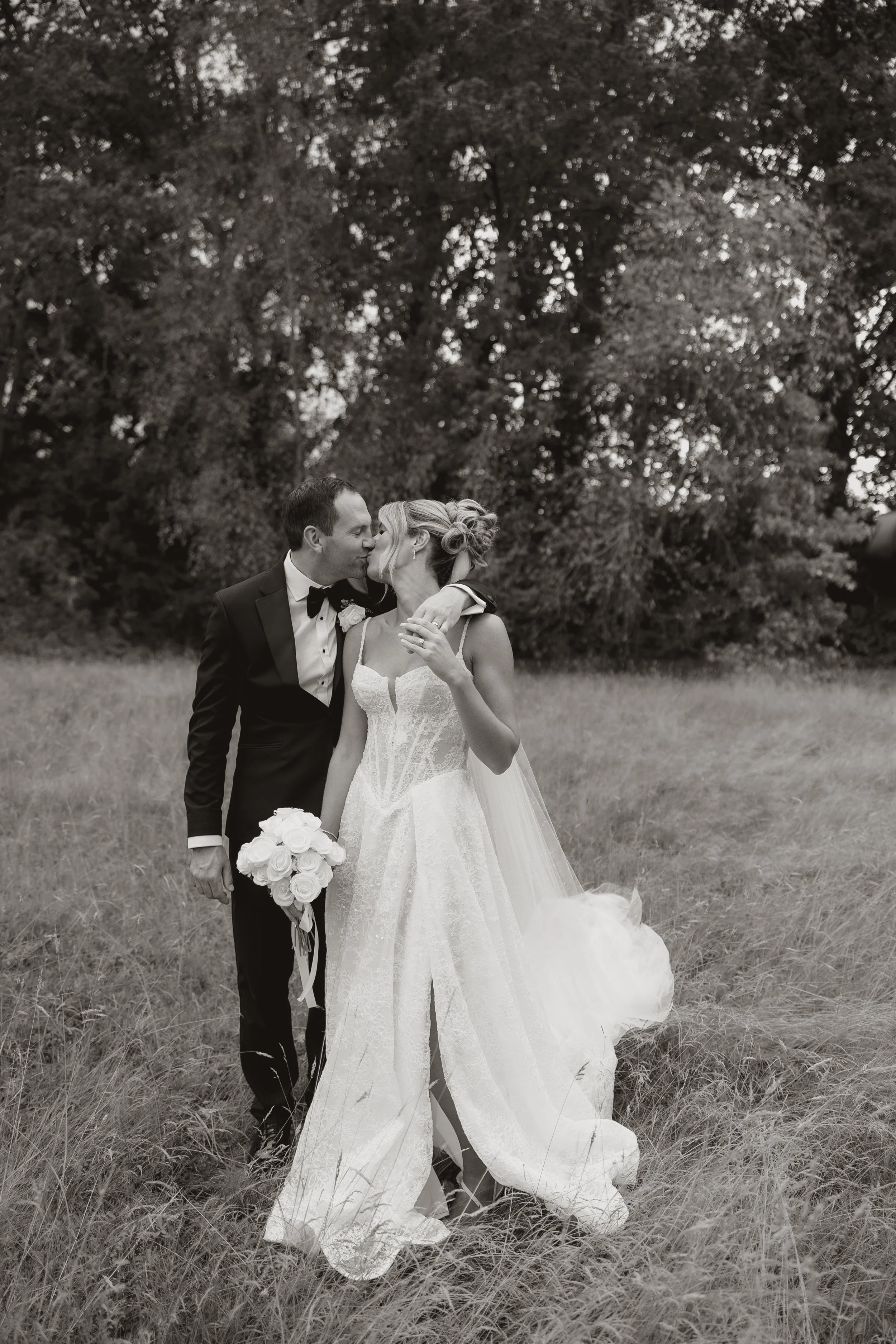 A black and white photo of a bride and groom kissing outdoors during their wedding. The groom is wearing a tuxedo holding a bouquet of white roses, and the bride is in a lace wedding dress with a veil, standing in a grassy field with trees in the background.