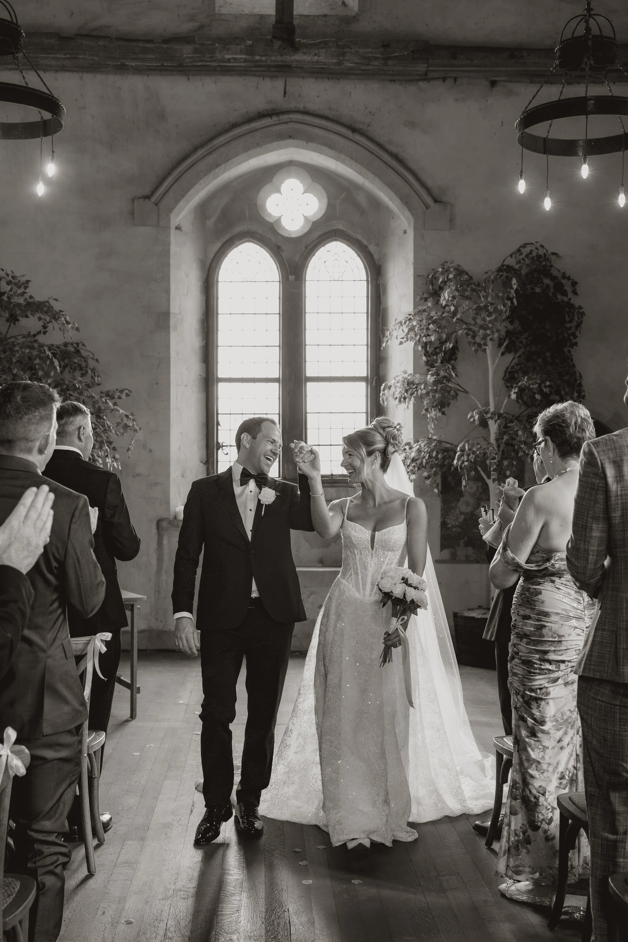 Black and white photo of a wedding ceremony inside a church with a bride and groom walking down the aisle, smiling and holding hands, surrounded by seated guests.