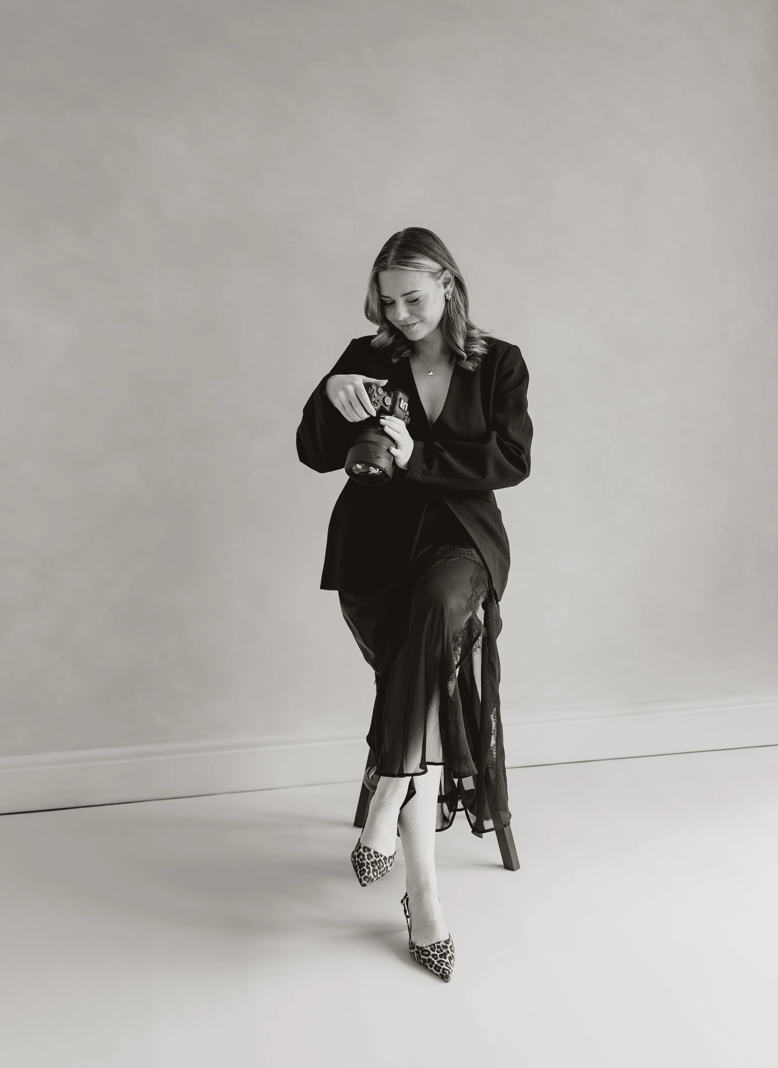 Black and white photo of a young woman sitting on a chair, looking at her camera, wearing a black dress, fishnet stockings, and leopard print heels, with a plain background.