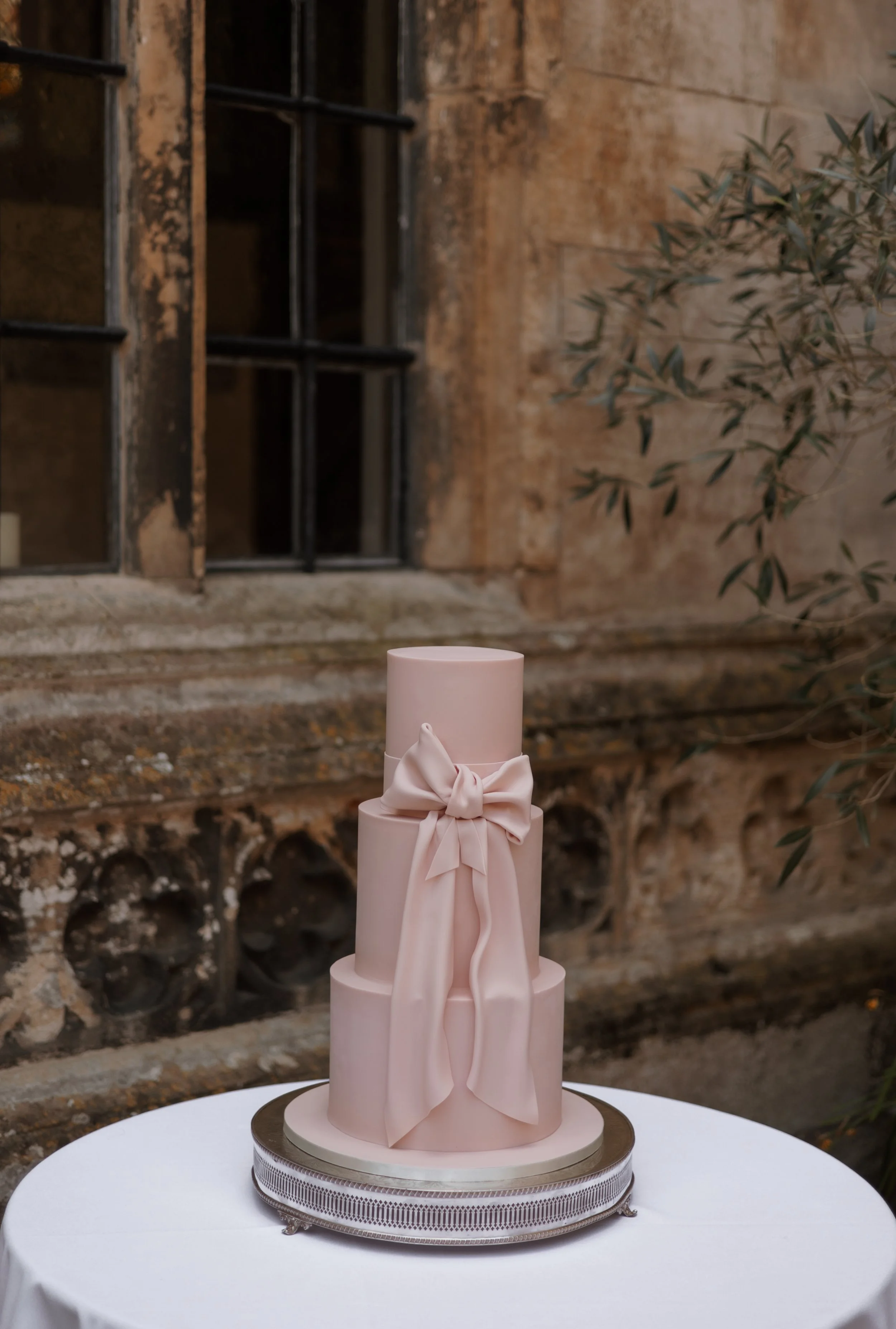 Three-tiered pink wedding cake with a bow decoration on the middle tier, placed on a silver cake stand on a white table, outdoors with a rustic brick wall and window in the background.