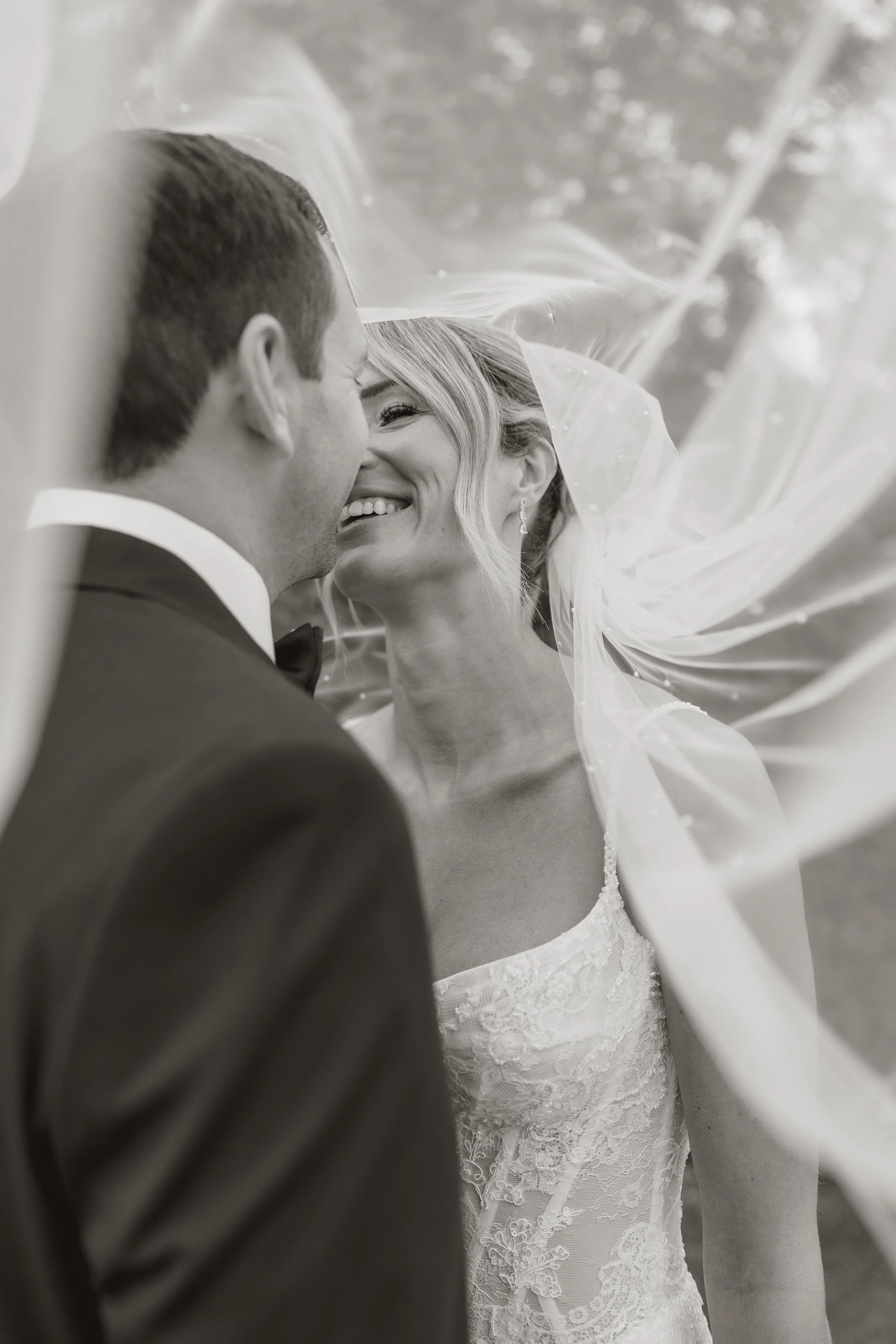 Black and white photograph of a bride and groom smiling and nuzzling under a sheer wedding veil, with the groom's back to the camera and the bride's face visible, wearing pearl earrings and a lace wedding gown.