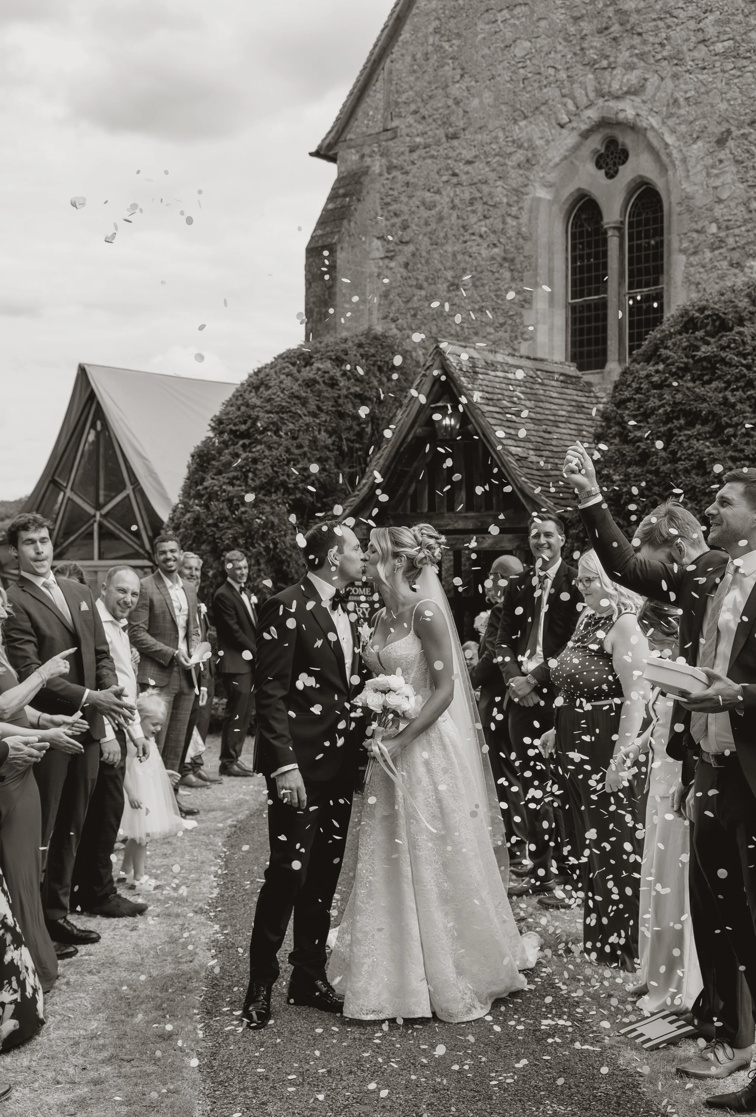 A black-and-white photo of a wedding celebration with a bride and groom kissing, surrounded by wedding guests throwing confetti outside a stone church