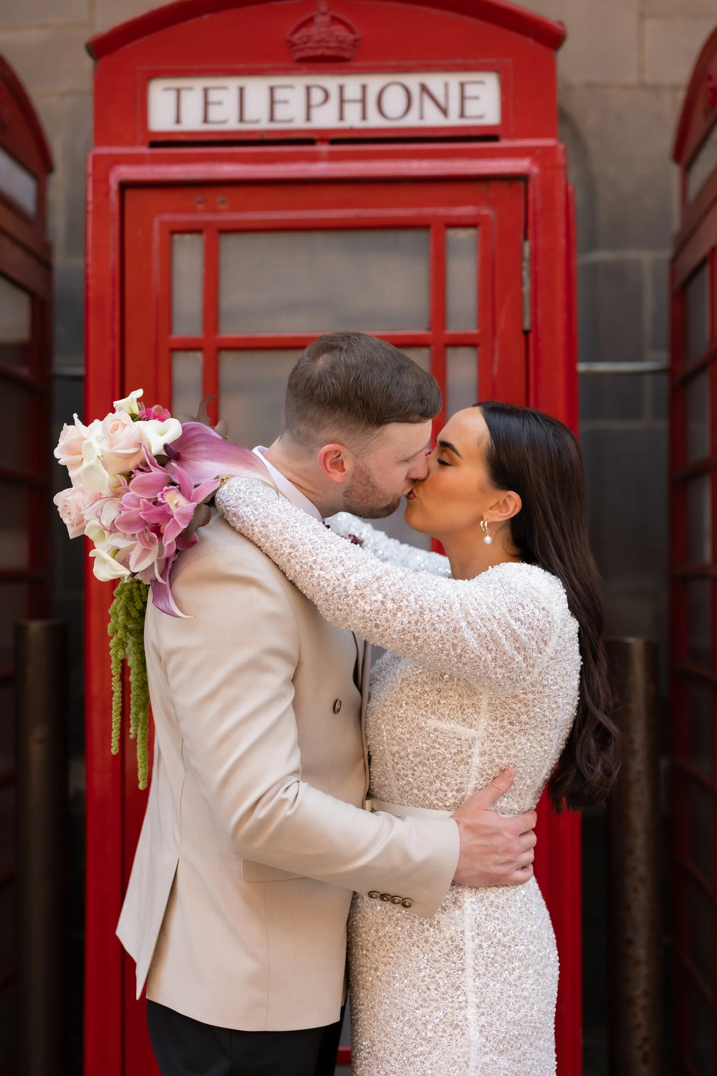 A wedding couple sharing a kiss in front of a red British telephone booth, with the bride holding a bouquet of pink and white flowers.