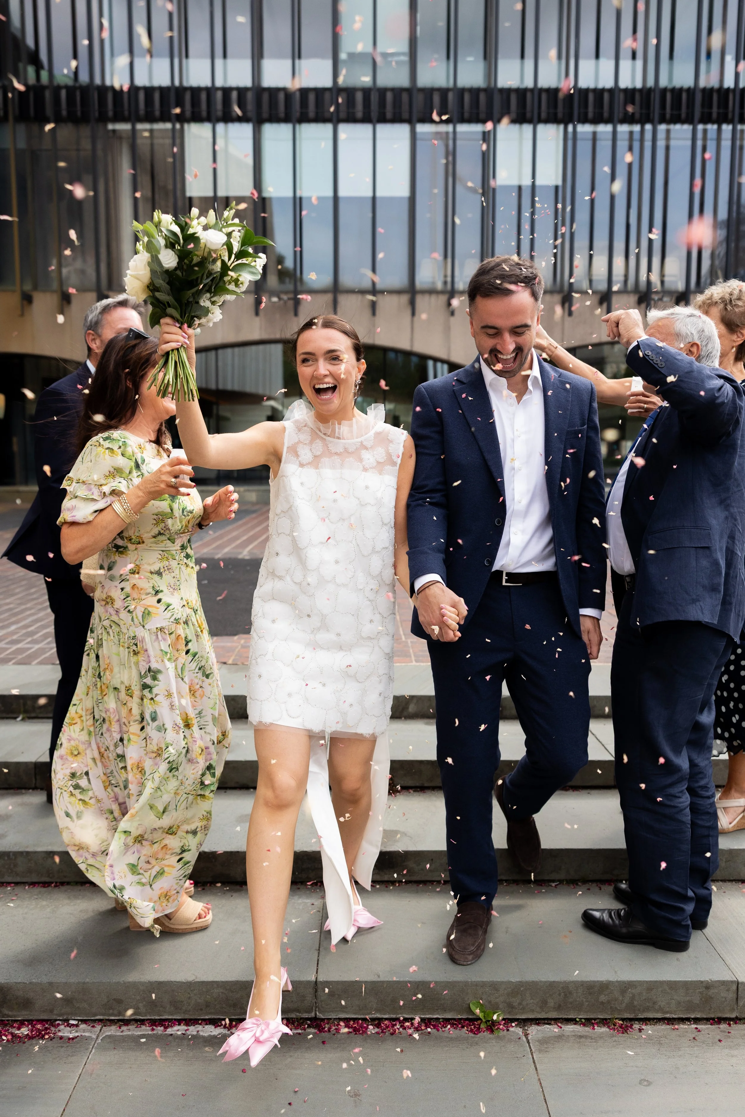 A wedding celebration outside a modern building with glass windows. The bride, in a white lace dress, holds a bouquet and smiles as she walks down the steps. A groom, in a navy suit, walks beside her holding her hand, both surrounded by happy guests. Pink flower petals are falling through the air.