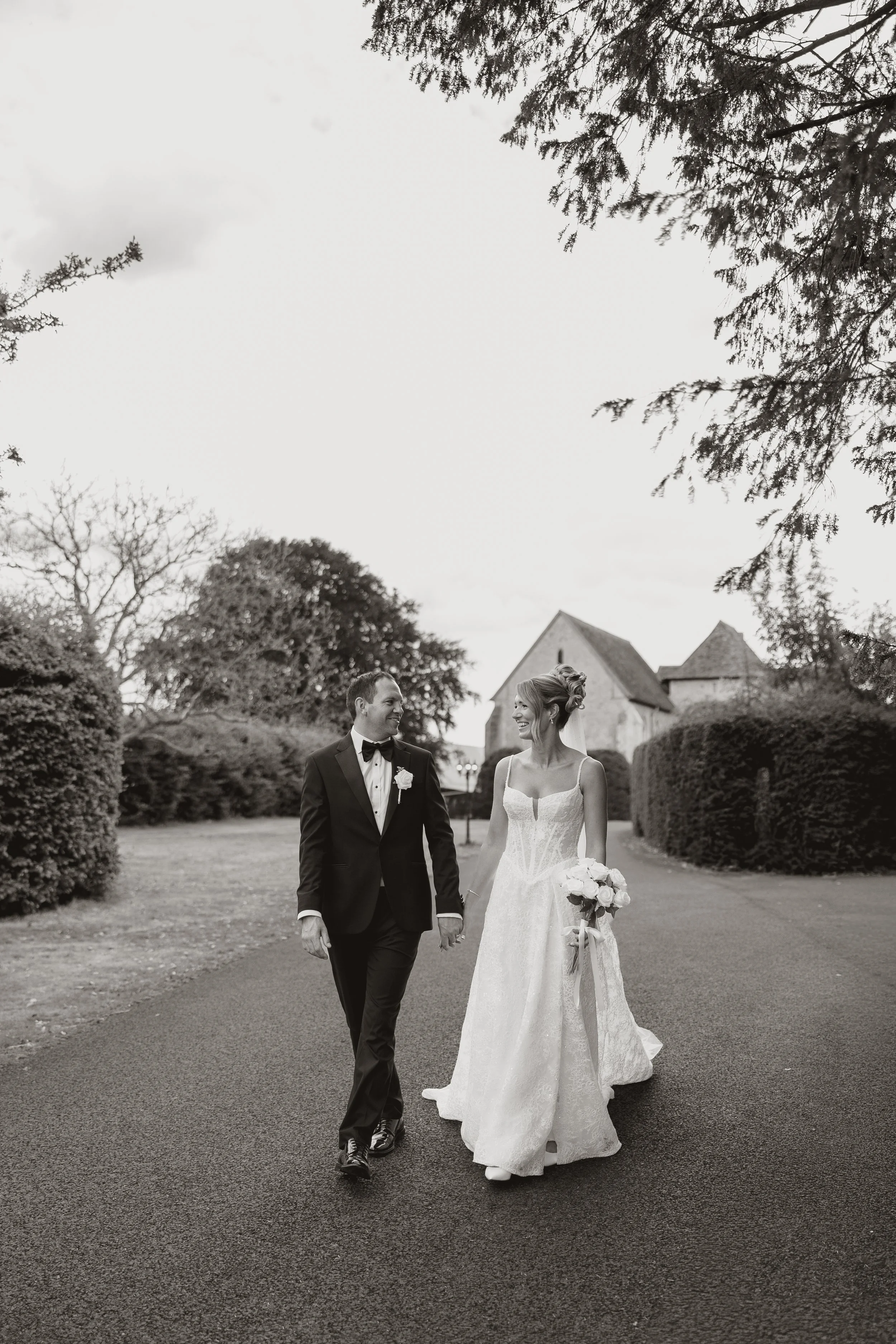 A bride and groom walk hand in hand outdoors, smiling at each other, in a garden with trees and a building in the background.