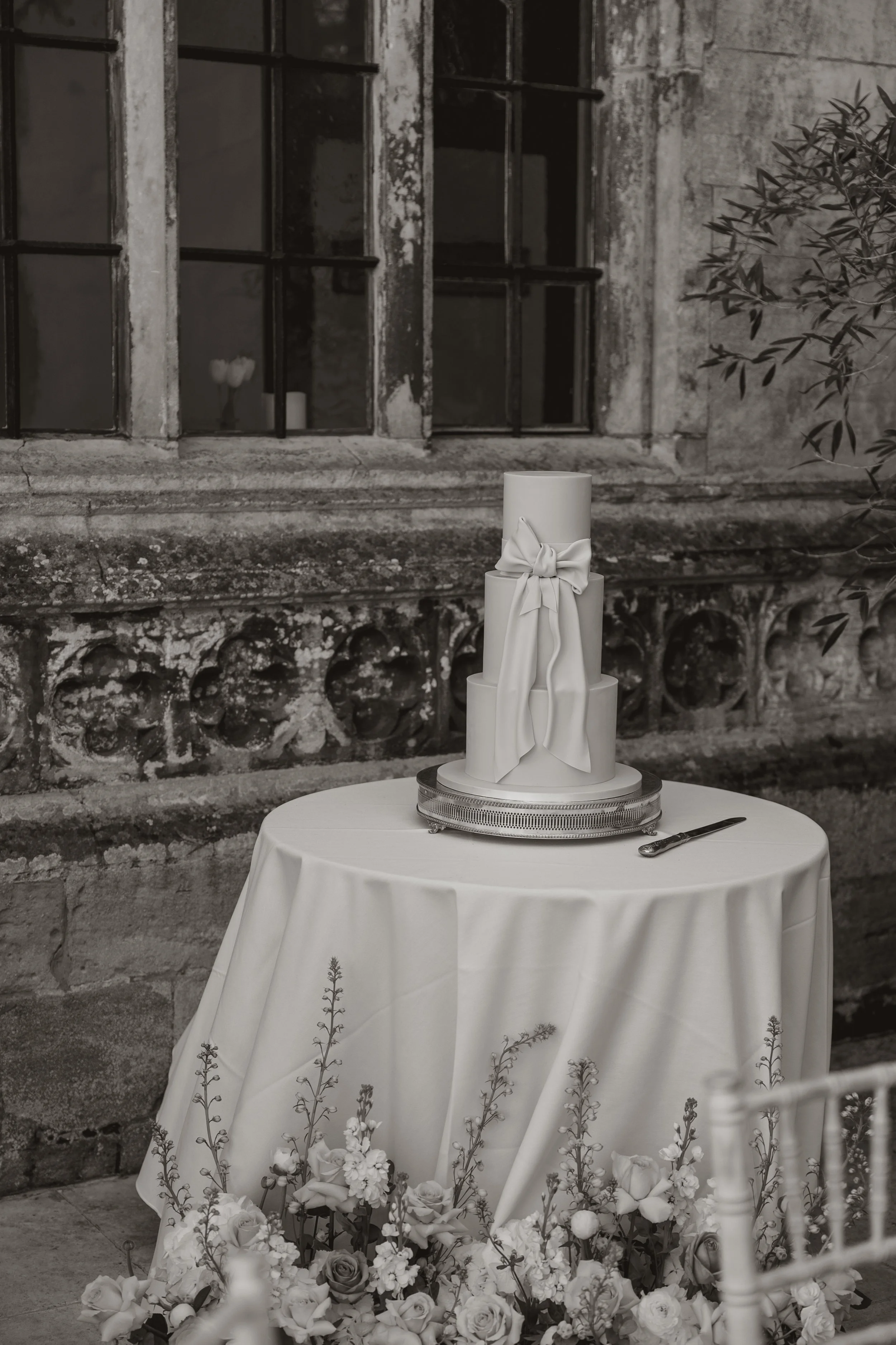 A tiered white wedding cake with a bow decoration on top, placed on a round table with a white tablecloth, surrounded by floral arrangements, against an old stone wall with window panes.
