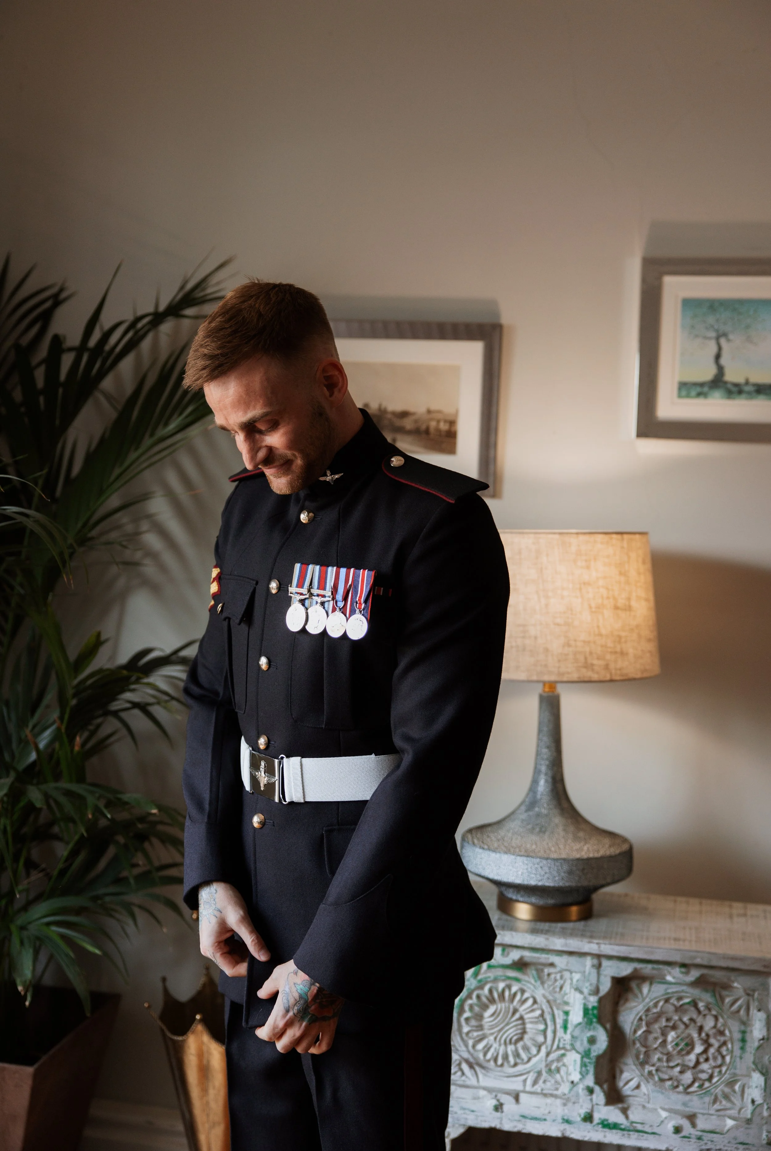 A man in a formal military uniform with medals stands indoors, smiling and looking downward, with a plant, paintings, and a table lamp in the background.