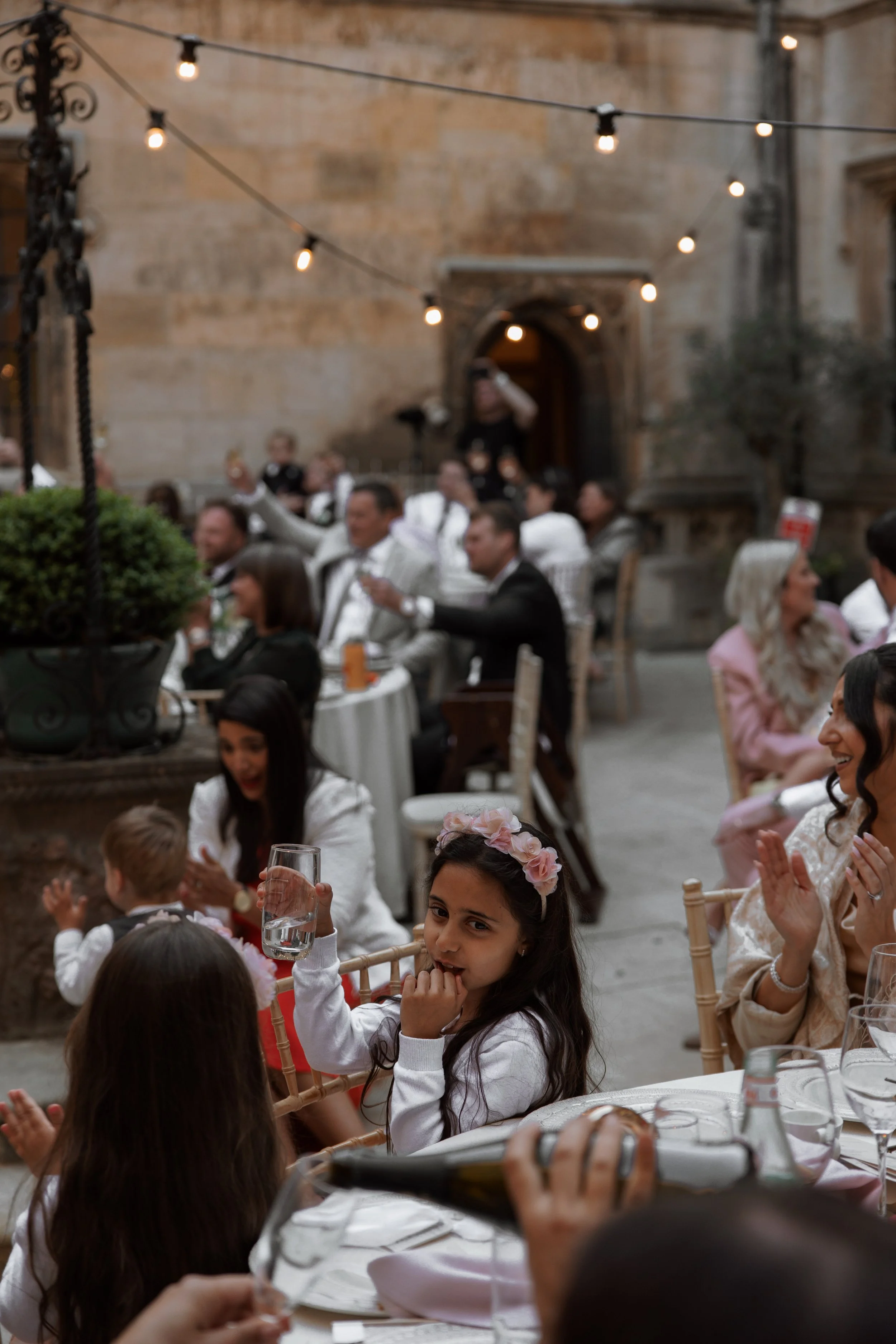 Wedding reception with children and adults at tables, children with flower crowns, adults in formal attire, outdoor setting with string lights and stone building in background.