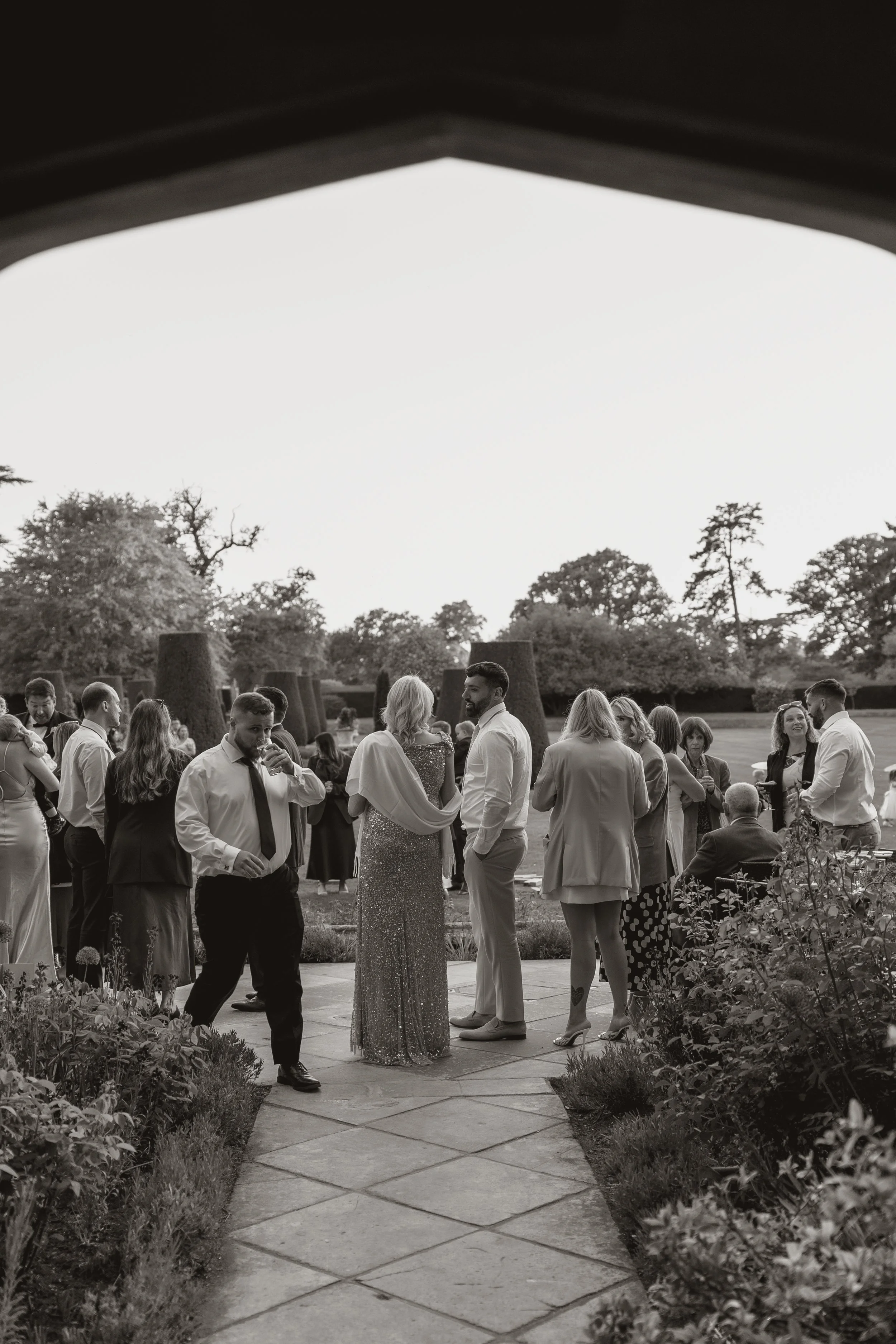 People mingling outdoors at a gathering, possibly a wedding, on a stone pathway surrounded by garden plants, with trees in the background, under an open sky.