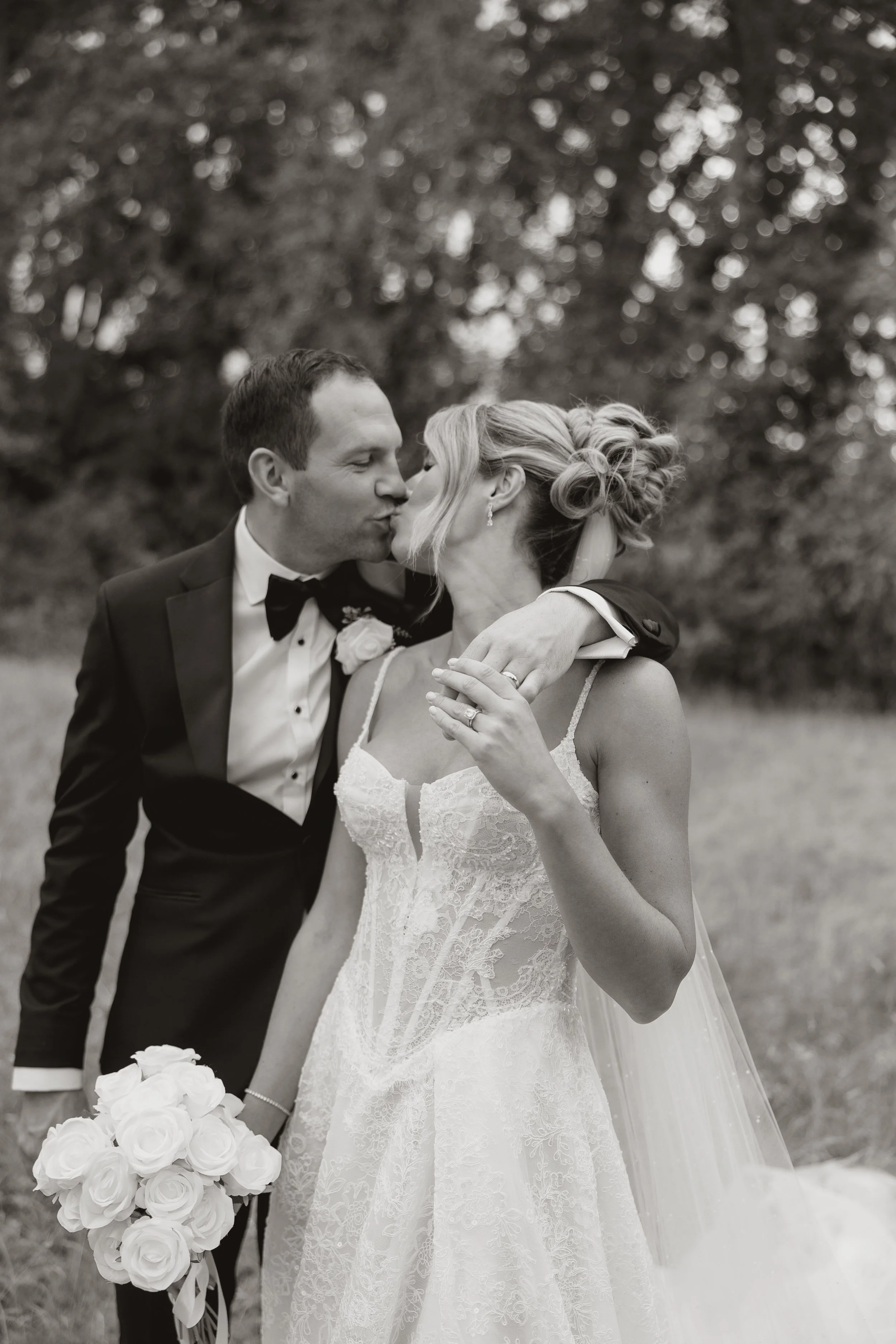 A black-and-white photograph of a bride and groom kissing outdoors, with the groom in a tuxedo and the bride in a lace wedding dress holding a bouquet of roses.