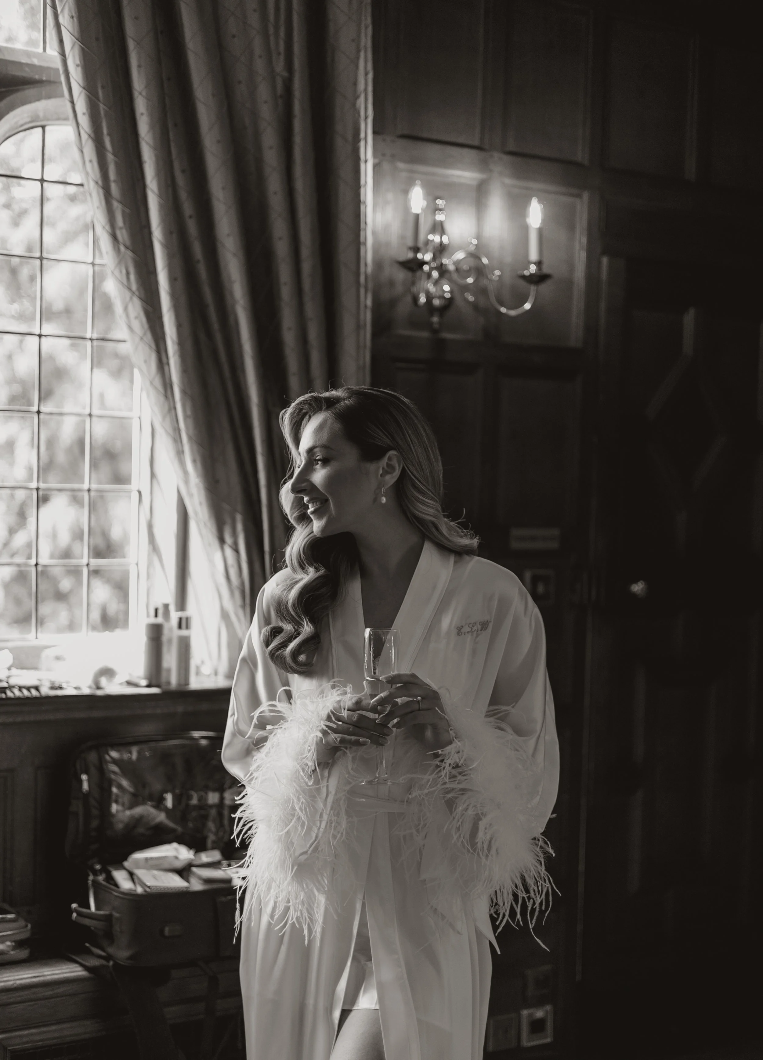 A woman in a silky robe with feathered trim holding a glass of champagne, standing near a window with drapes and a chandelier in the background.