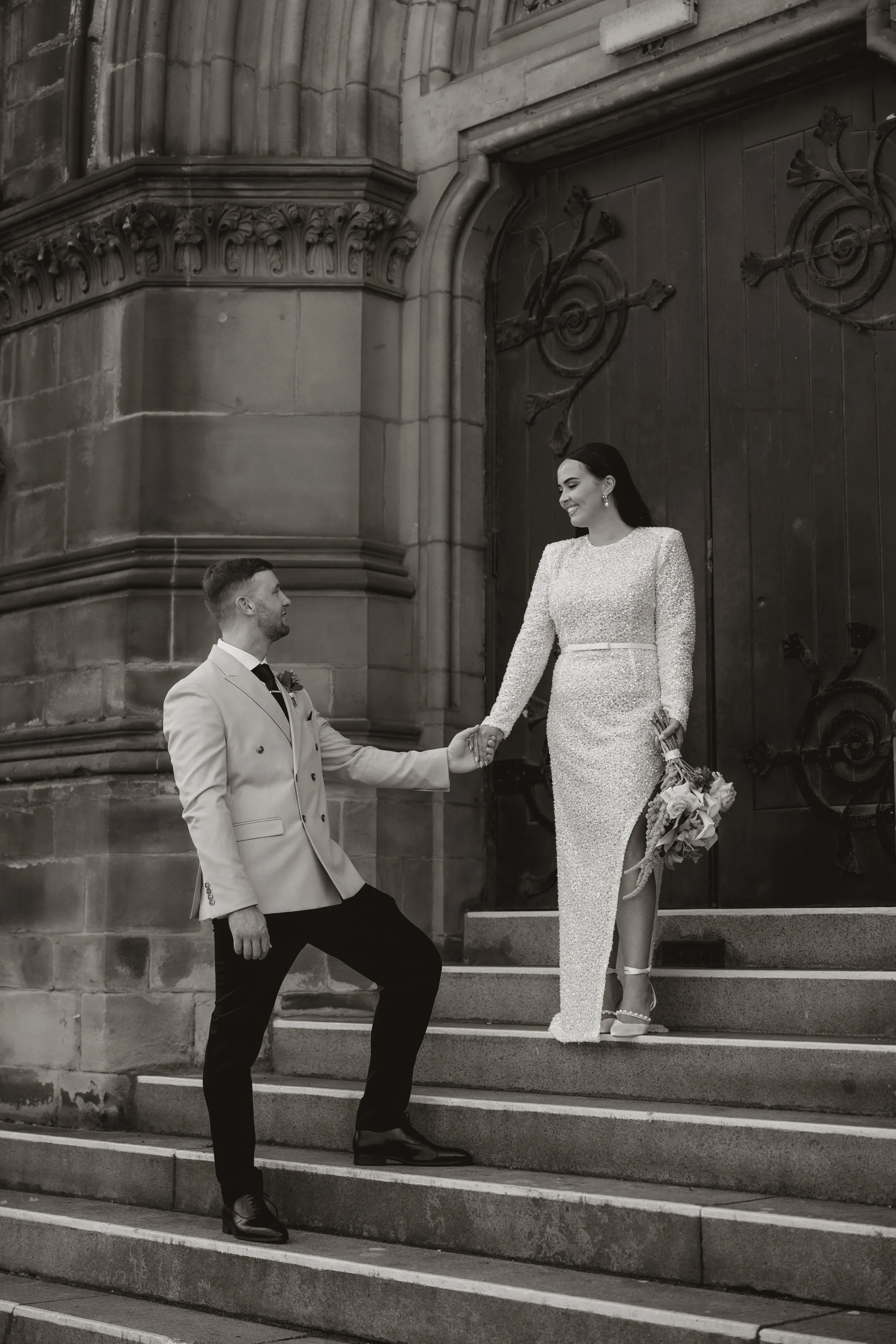 A black and white photo of a man kneeling on staircase steps, holding hands with a woman standing on the steps. The woman is dressed in a long, sparkly gown and holding a bouquet, smiling at the man. They are in front of an ornate building entrance w