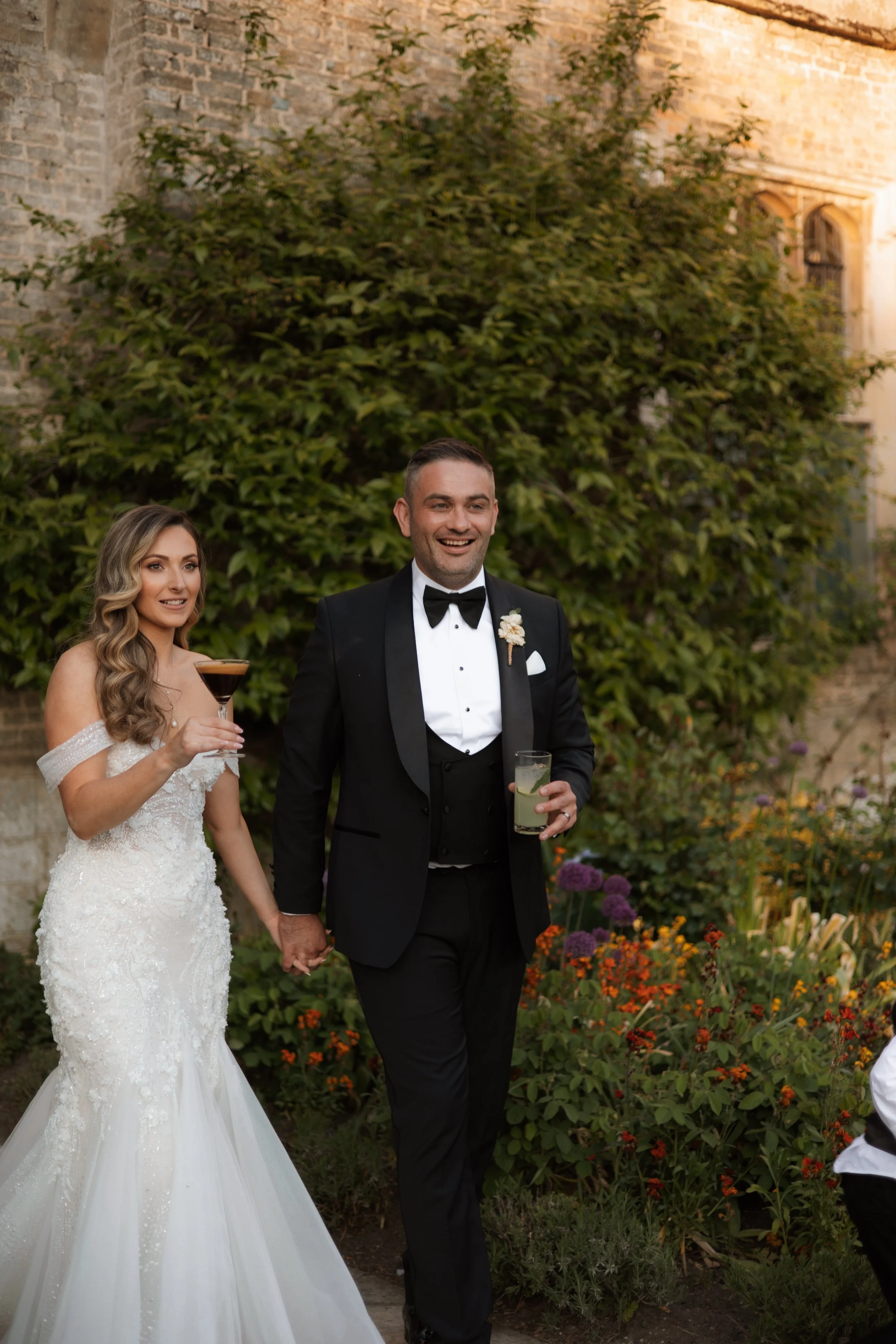 A newlywed couple holding hands outdoors during their wedding celebration, with the bride holding a cocktail and the groom holding a drink, standing in front of a lush greenery and brick building.