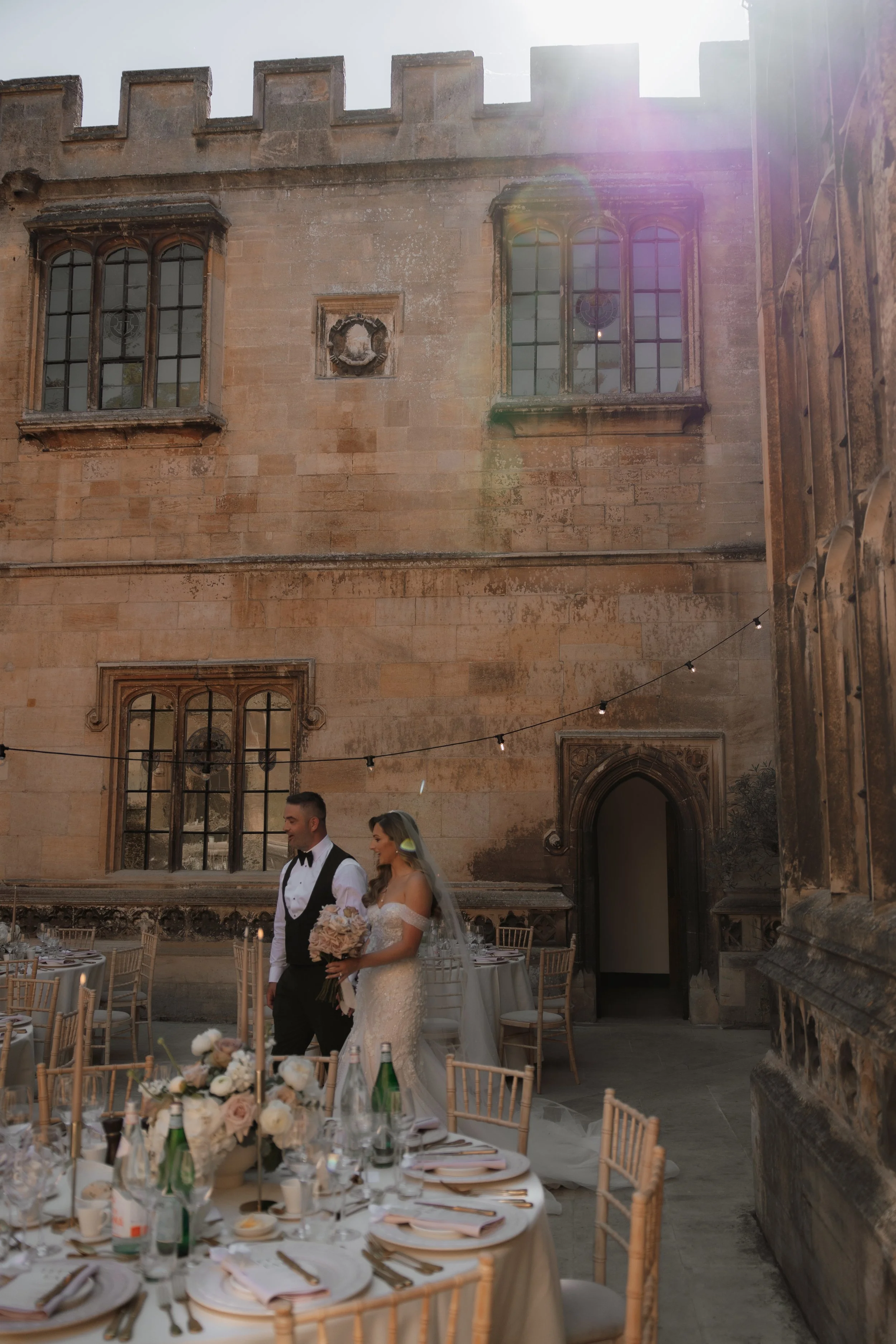 A bride and groom at a wedding reception in an outdoor courtyard with historic stone walls, large gothic windows, and decorated tables with floral centerpieces.
