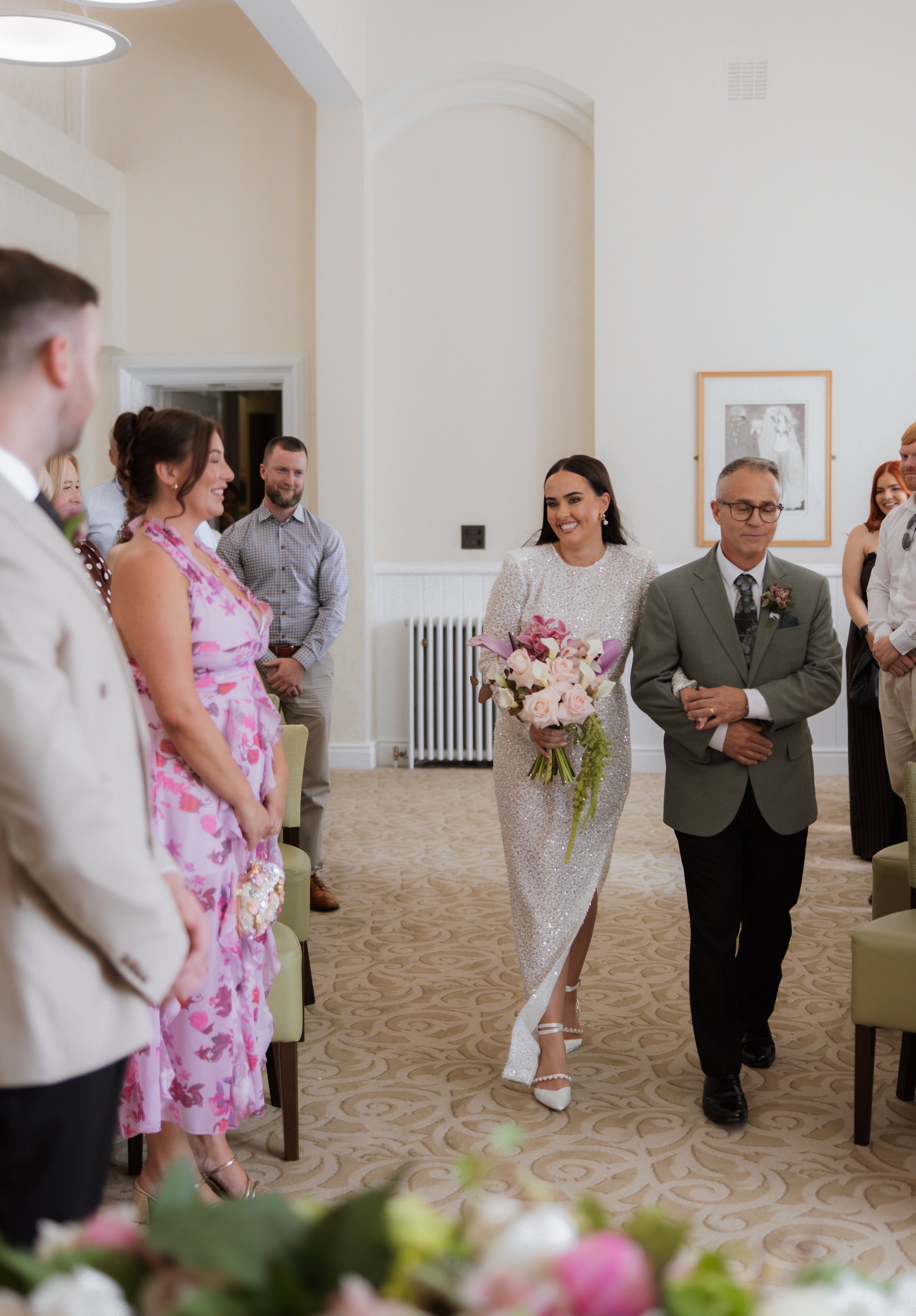 A bride in a white, sparkly wedding dress with floral bouquet walking down an aisle, accompanied by a groom in a grey suit, surrounded by smiling guests in a warmly lit room with cream-colored walls and framed artwork.