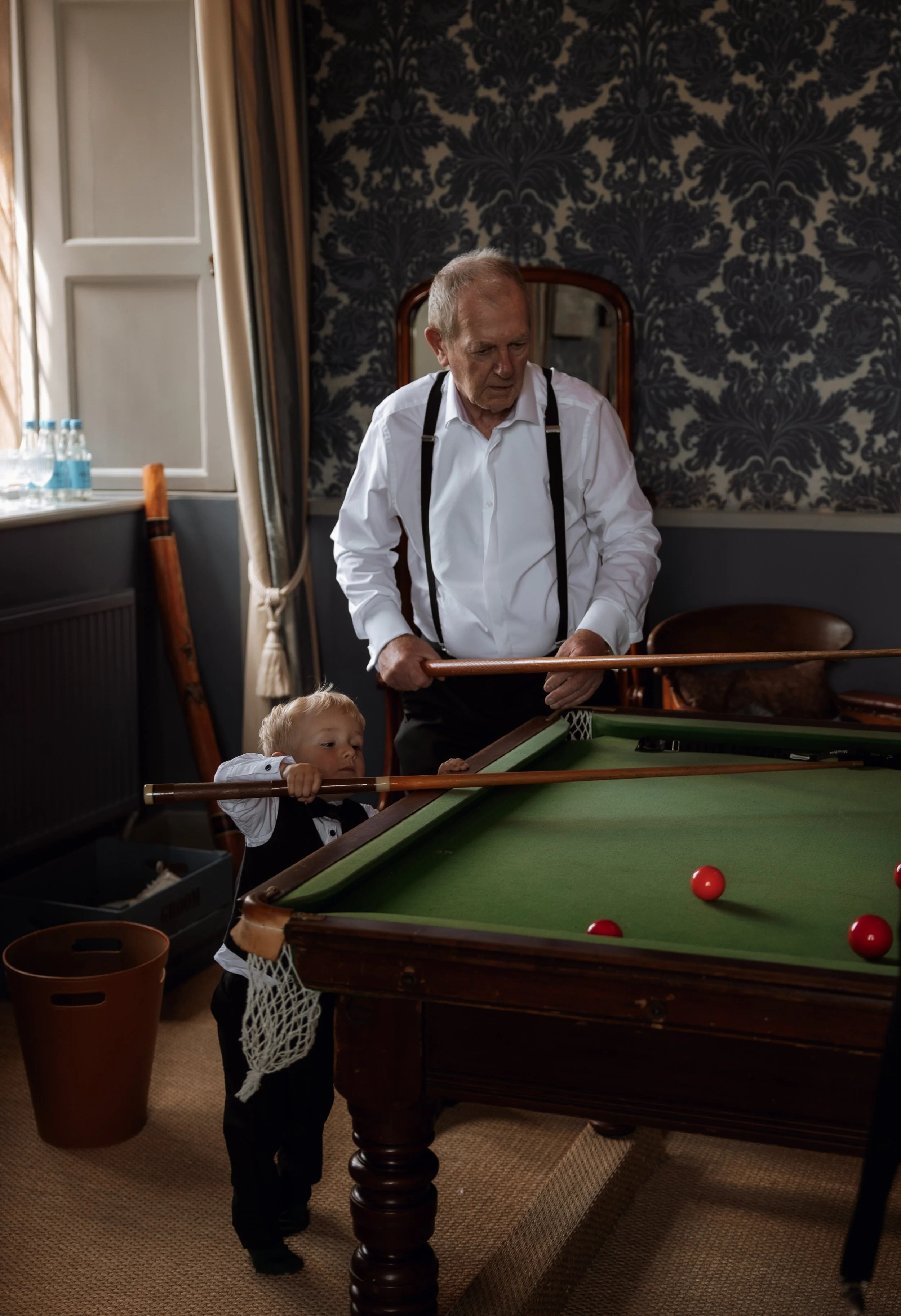 An elderly man in a white shirt and suspenders is playing billiards with a young boy, who is holding a cue stick and aiming at the balls on the pool table in a dimly lit room with dark patterned wallpaper and a window with curtains.