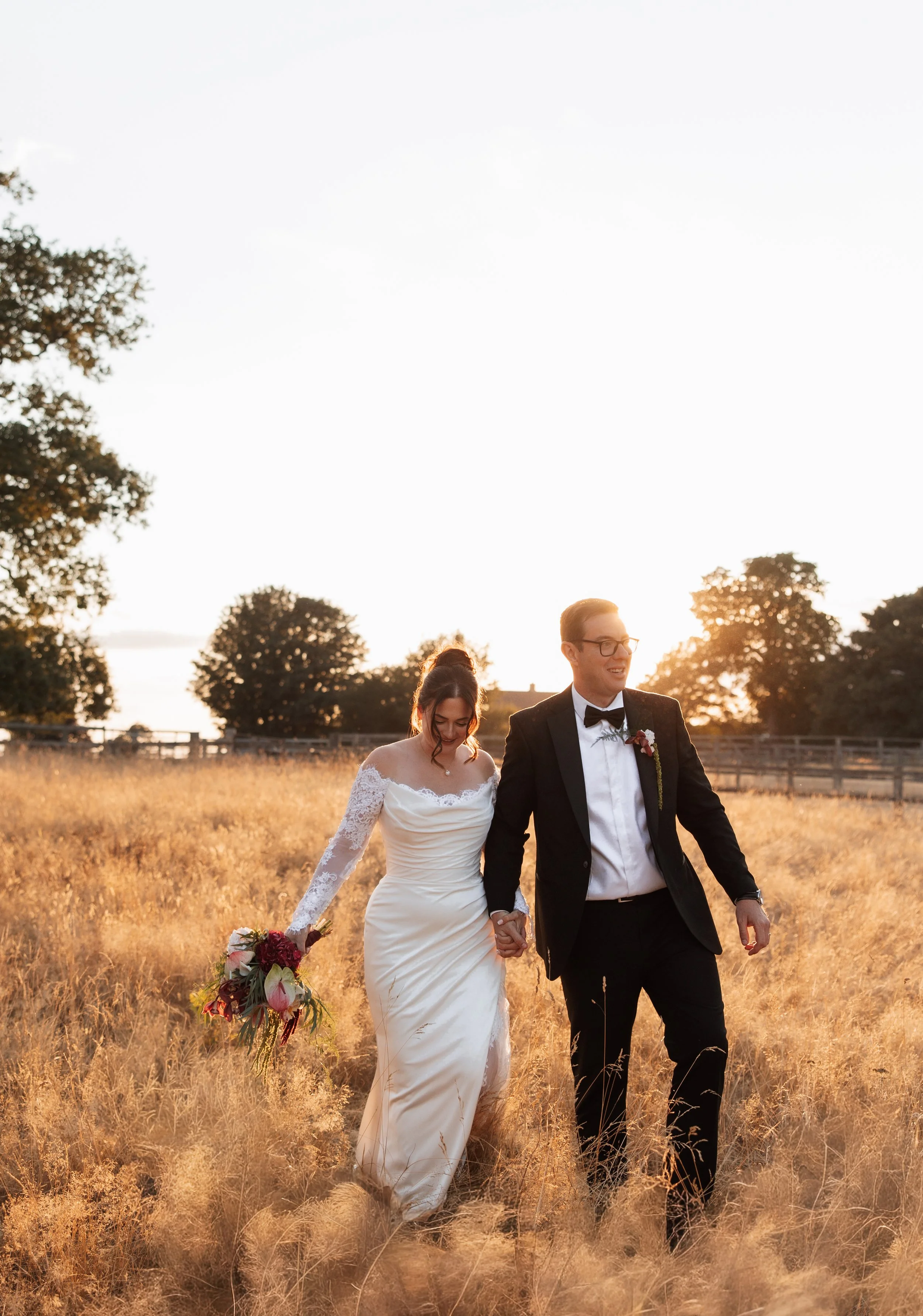 A bride and groom walking hand in hand through a field of tall, golden grass during sunset, with trees in the background and a bright sky above.