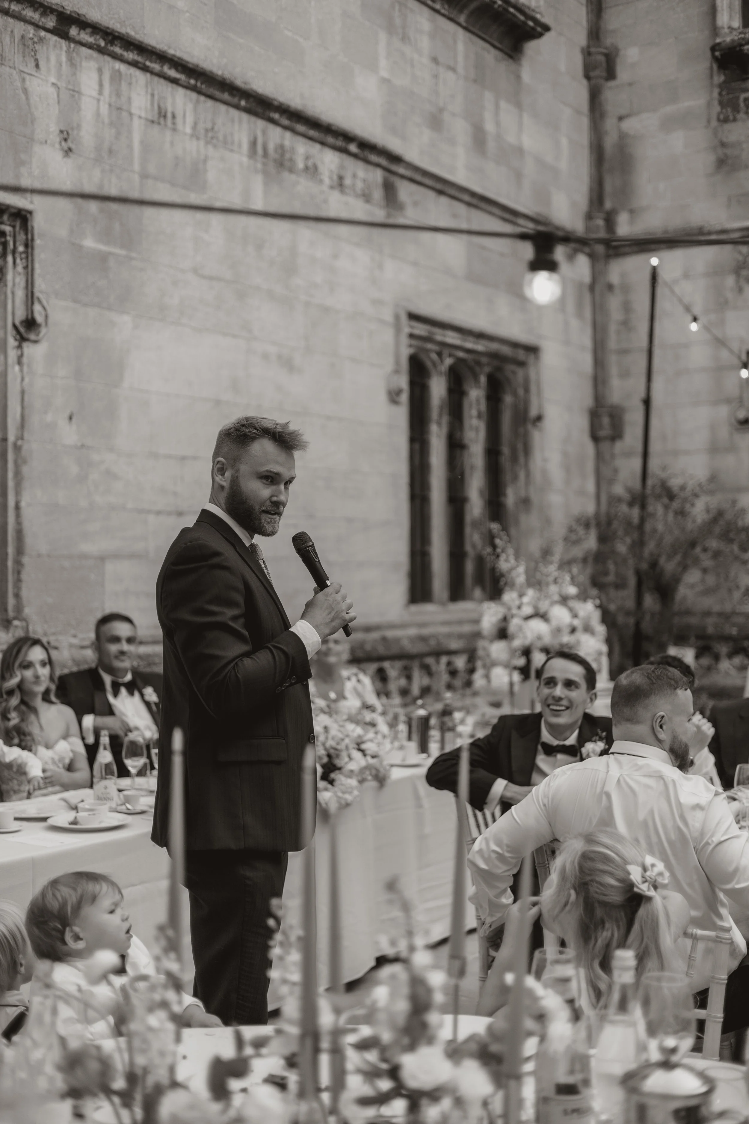 A man giving a speech at a wedding reception, holding a microphone, while guests sit at a decorated table with floral arrangements and candles.