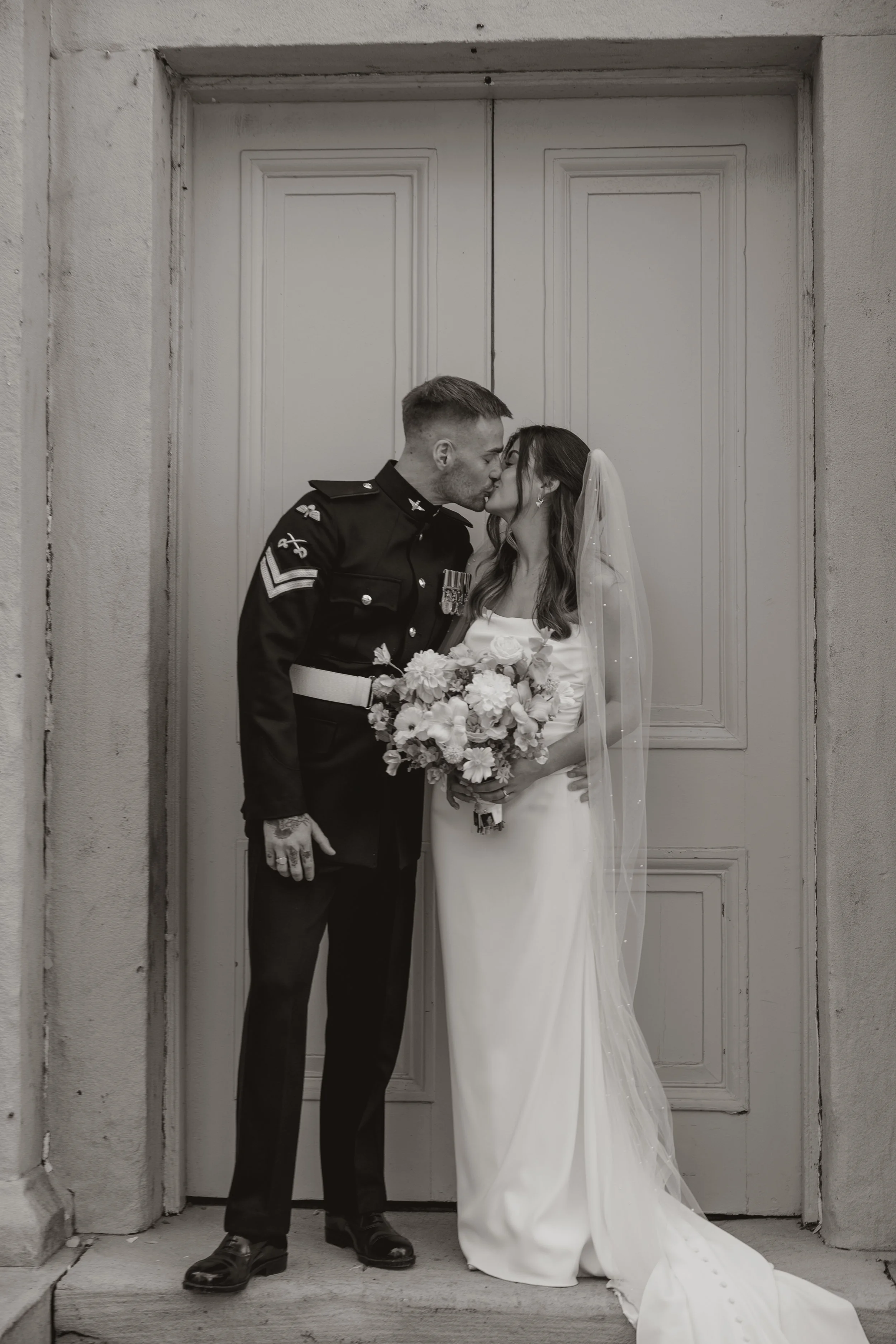 A black and white photo of a bride and groom sharing a kiss in front of a door. The groom is in a military uniform and the bride in a wedding dress holding a bouquet of flowers.