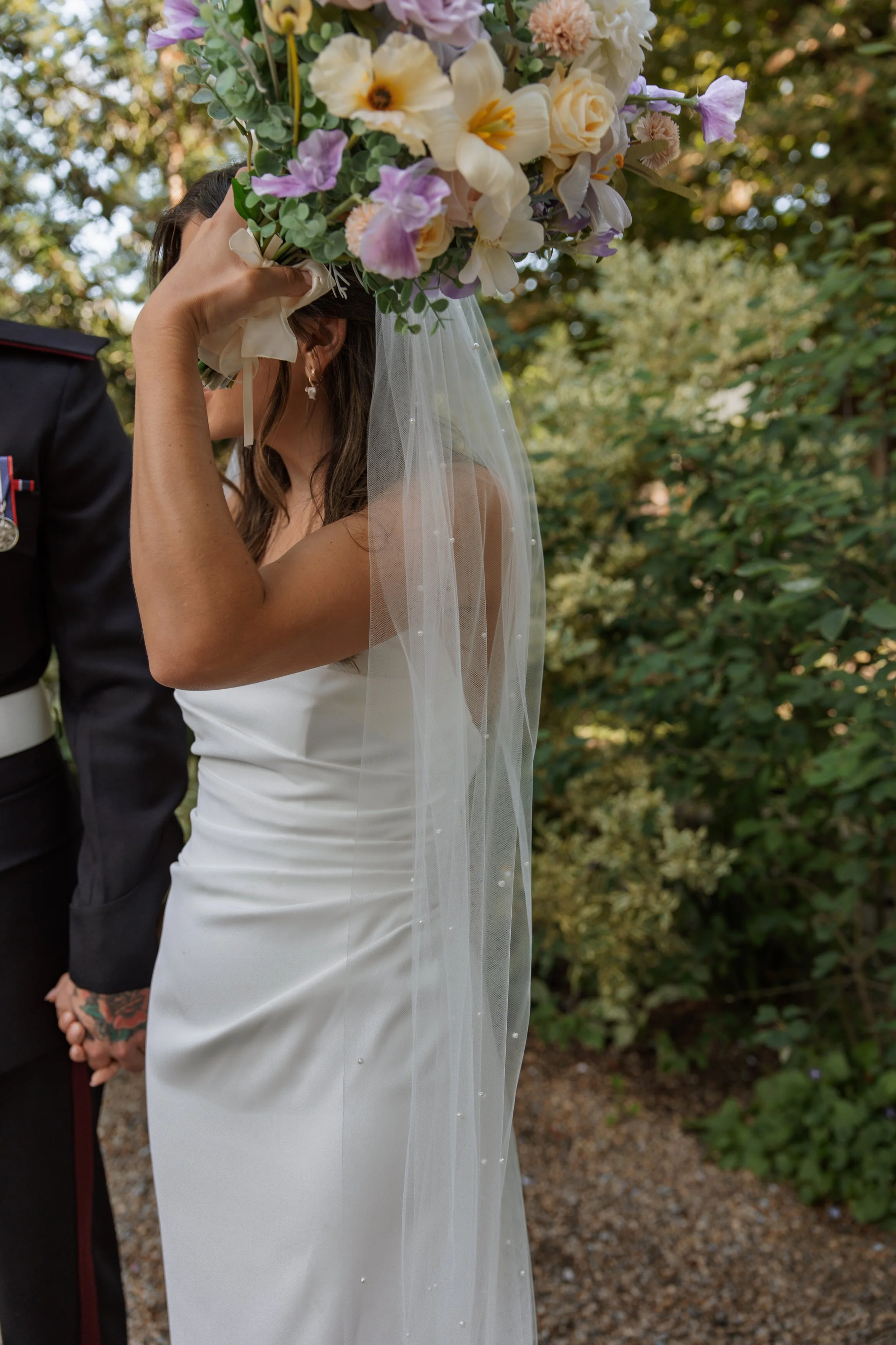 A bride holding a large bouquet of flowers over her head during a wedding ceremony outdoors, with greenery and trees in the background.