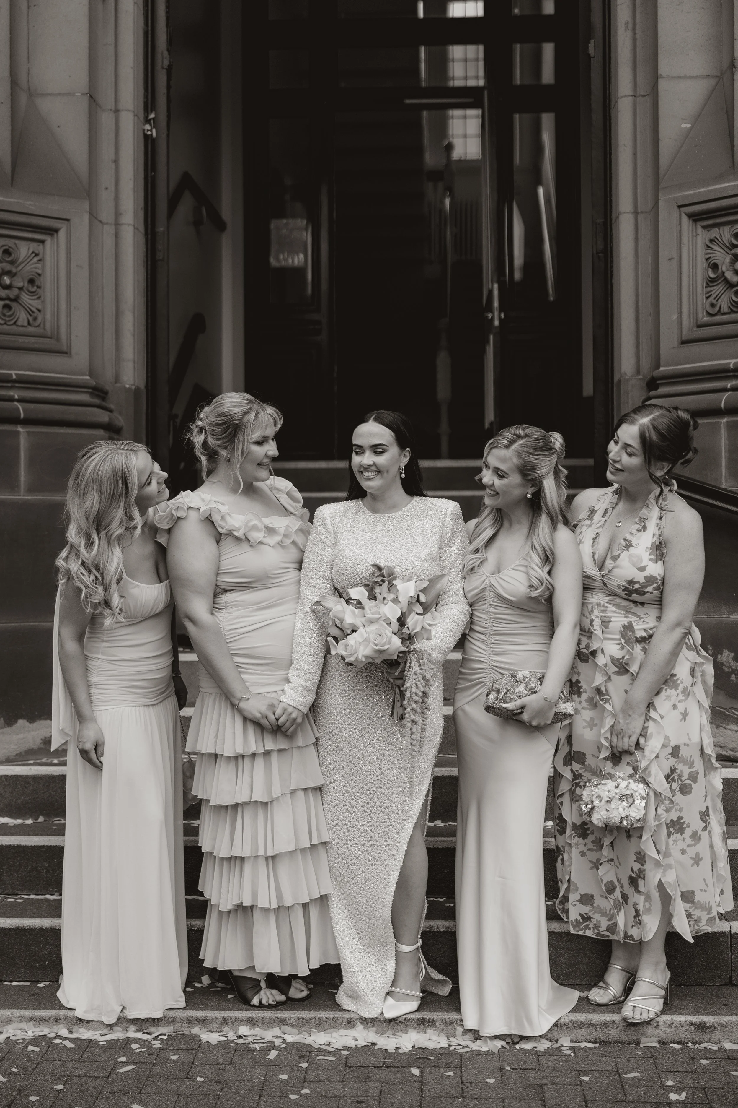 A group of six women dressed in formal attire, standing on steps in front of a building, with a bride holding a bouquet of flowers at the center, all smiling and looking at each other.